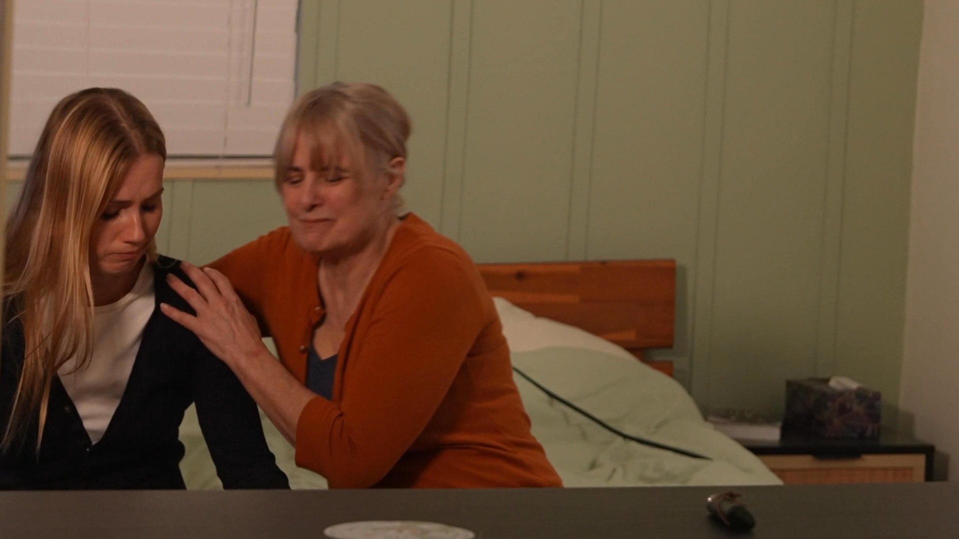 An older woman offers guidance and emotional reassurance to a younger woman sitting on a bed in a bedroom. The interaction centers on empathetic dialogue and physical comfort to address distress.