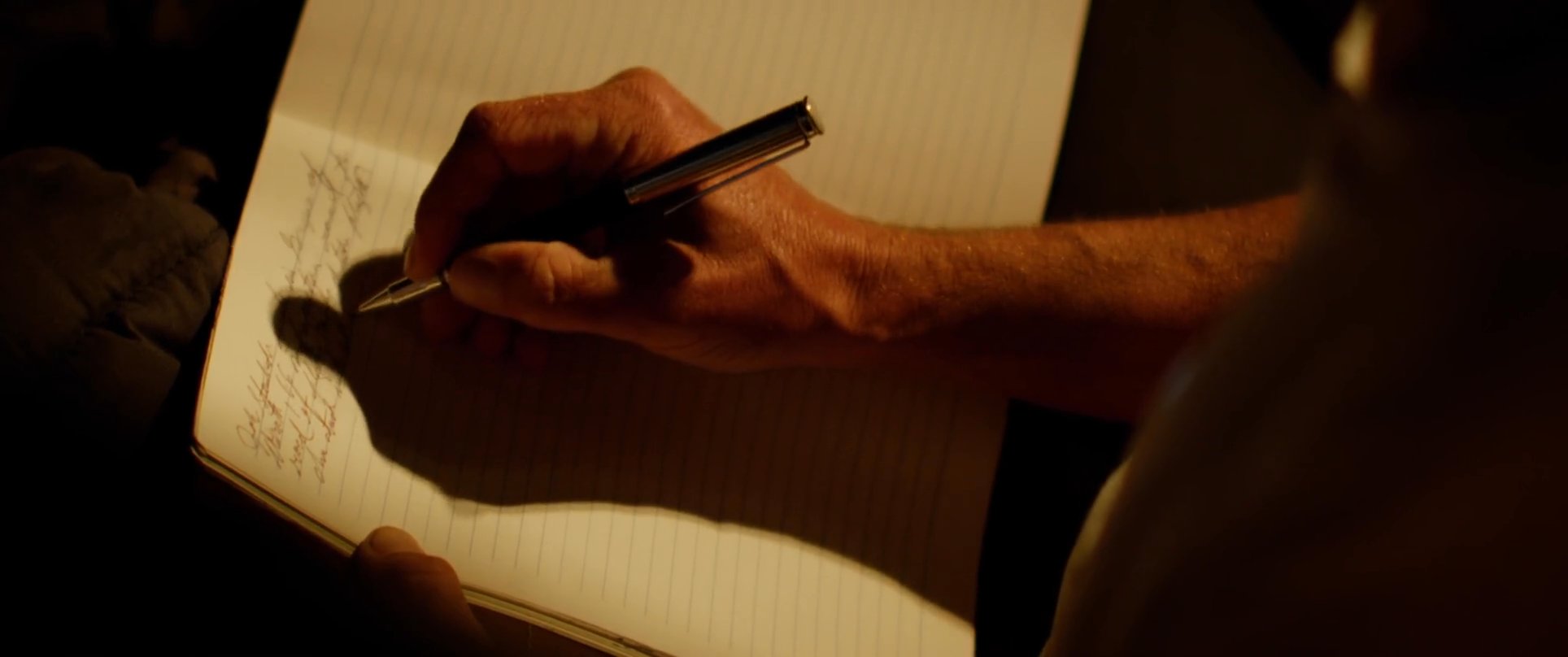 A man sits in the back of a truck illuminated by a lantern, writing thoughtfully in a notebook. He pauses to look into the distance, capturing a moment of quiet introspection.