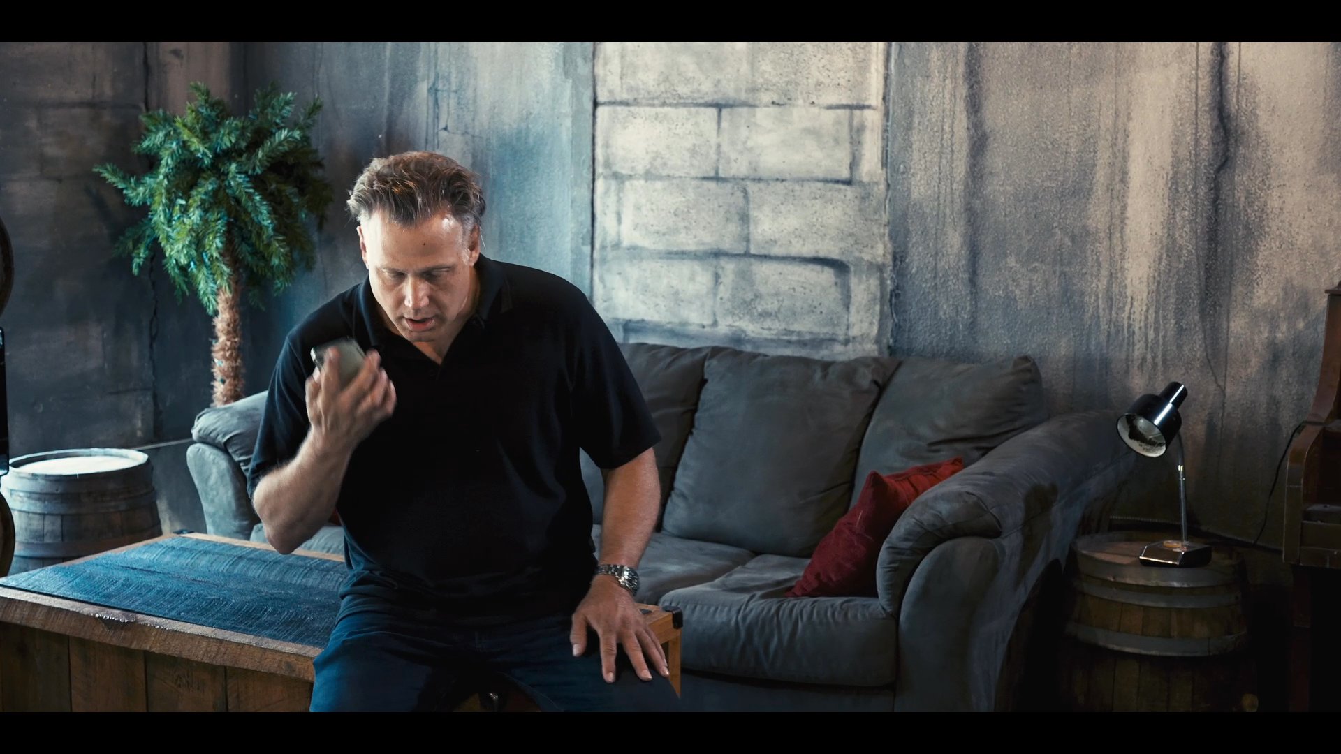 Three men engage in a professional discussion at a metal bar before one individual moves away to handle a private phone call. The scene transitions to focus on the individual as he conducts a serious conversation in a dimly lit, contemporary living space.