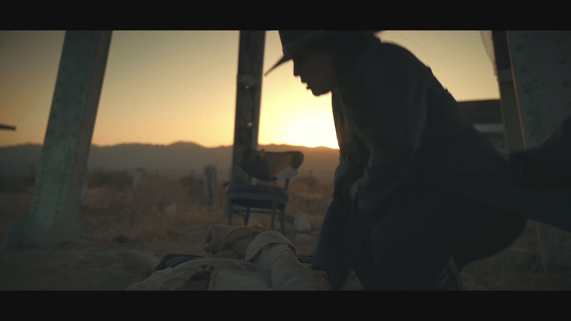 A black-clad, hat-wearing gunfighter stands beside a fallen person in a dusty frontier setting at sunset. The gunfighter crouches to handle the person’s hand and examines a small object as a dry, darkly comic exchange unfolds.