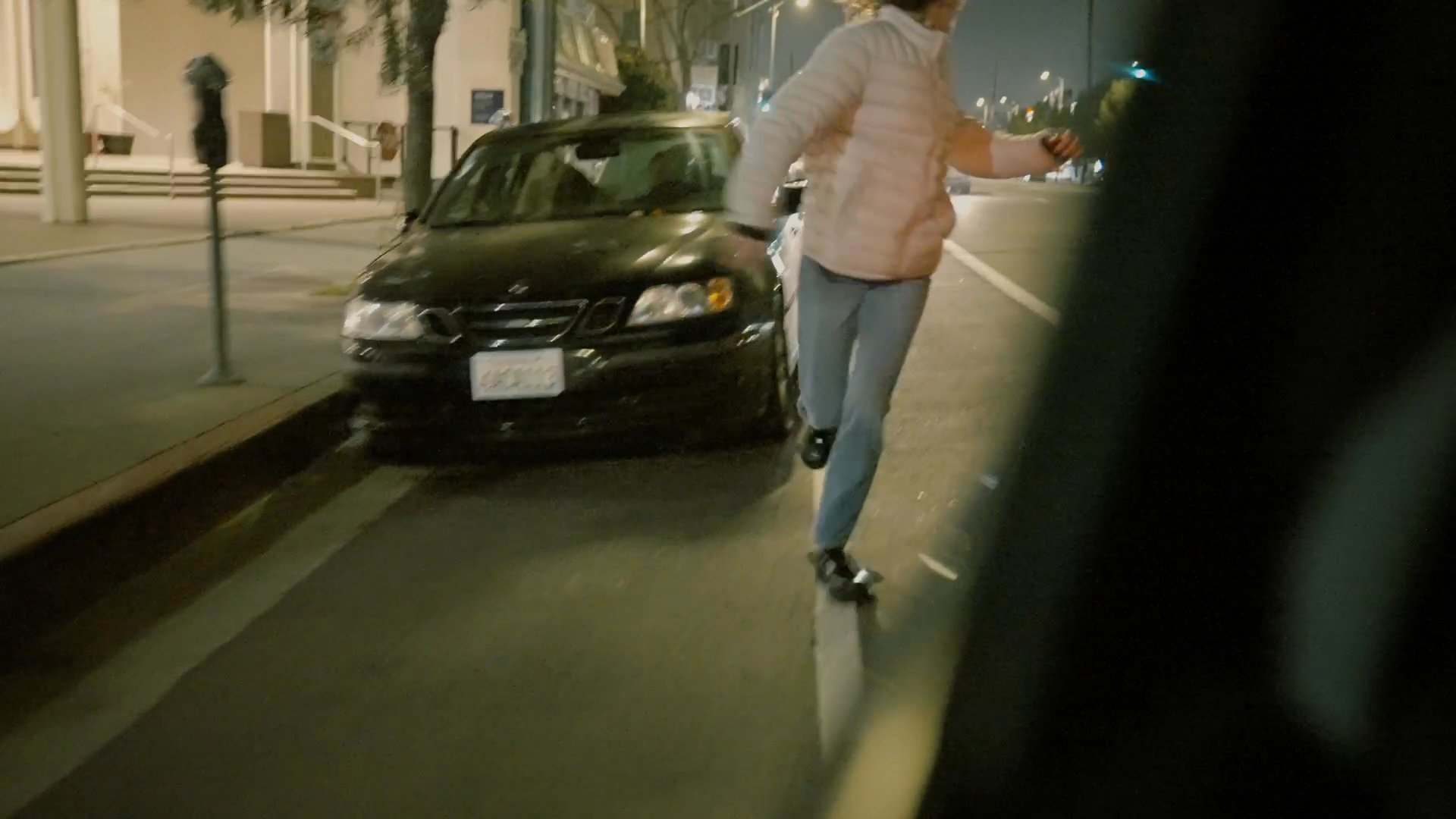 A woman runs through a city intersection at night while two men observe from inside a nearby vehicle.