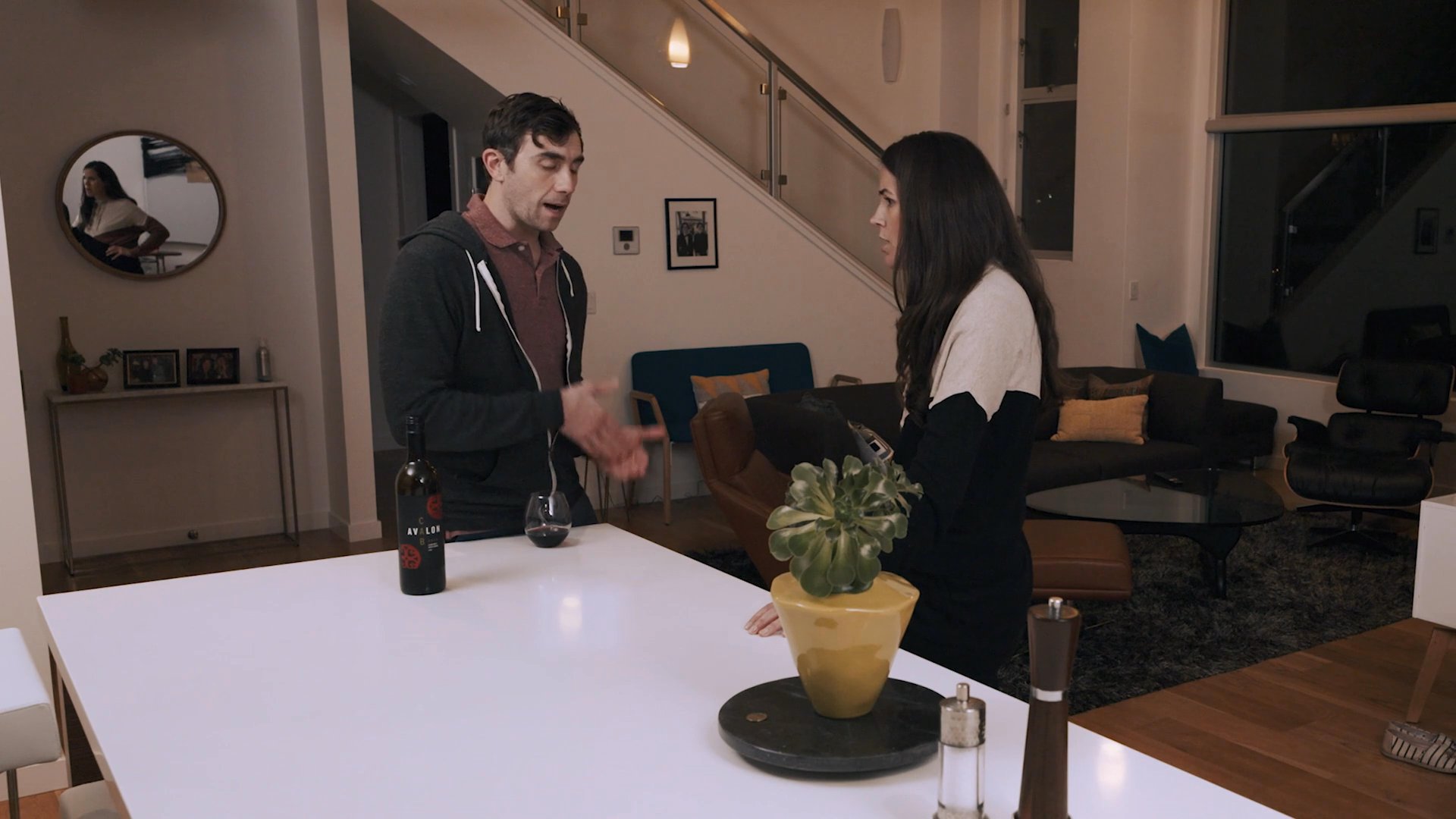 A man and woman engage in a serious, focused conversation while standing across from each other at a kitchen island in a residential home.