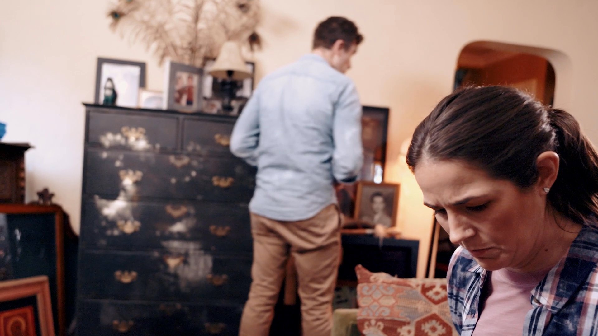 A man and a woman navigate a moment of interpersonal strain within a home living room setting. Their hesitant movements and focused expressions underscore a quiet, underlying conflict.