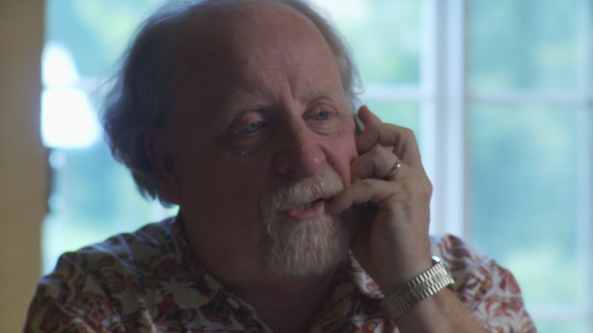An older man with a beard sits indoors by a window, listening and speaking during a focused telephone conversation. His facial expressions convey a sense of serious engagement as he listens to the caller.