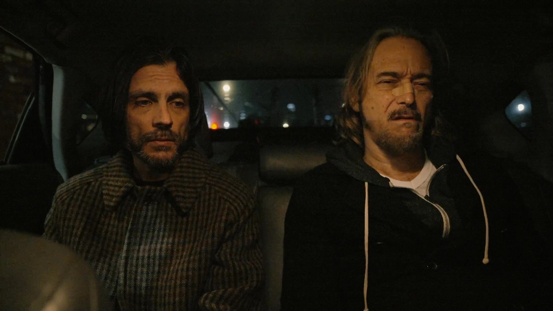 Two men observe a parking garage from inside a parked vehicle at night. While one monitors the exterior, the other checks his smartphone.