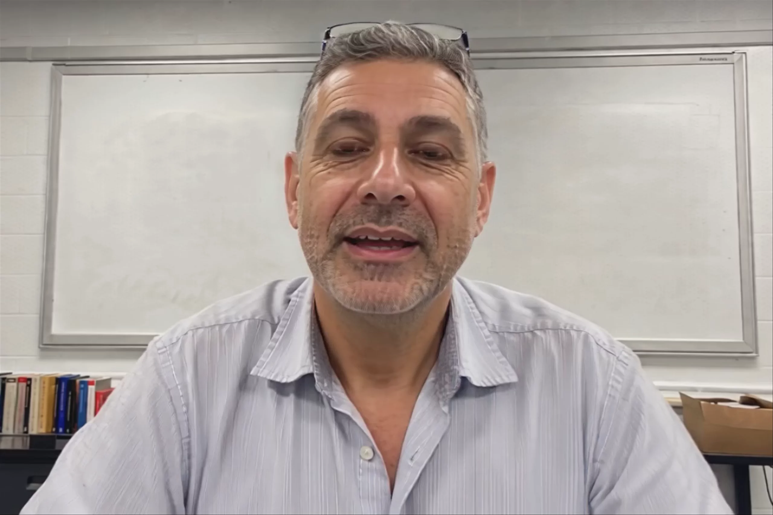 A widescreen 1080x720 seated talking-head video of a teacher at a desk in front of a whiteboard, warmly explaining a math concept to camera. Generated from a still image and an audio track using Hedra Character 3.
