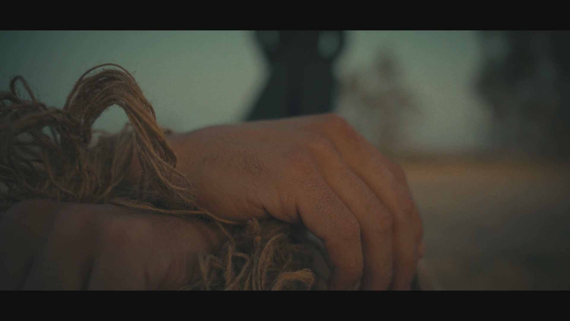 A black-clad figure in a wide-brimmed hat walks beneath a weathered timber structure at sunset. The sequence cuts to a close-up of a hand resting on a brown, rope-covered surface as upbeat music plays and someone says, “Cheers.”