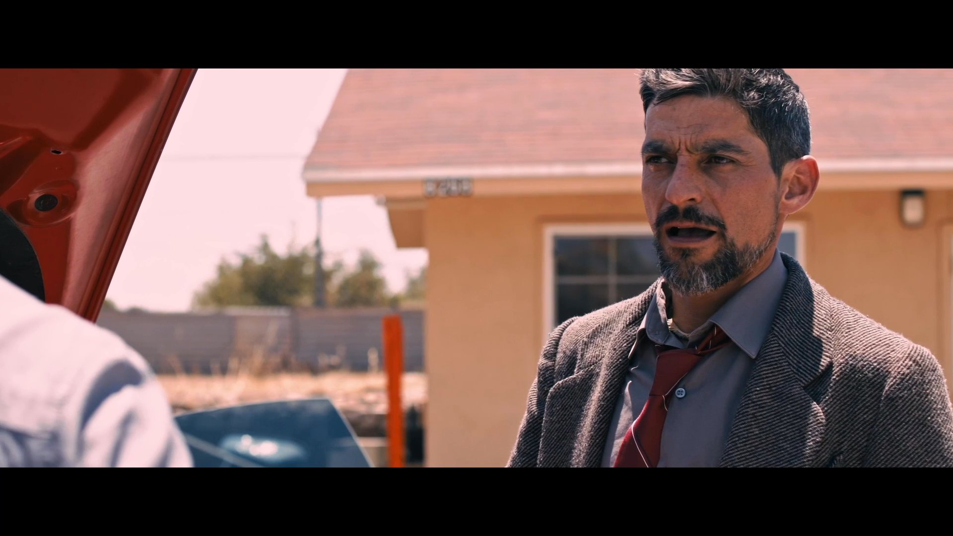 An adult male in business attire engages in a conversation with two younger men in a parking lot, while a police patrol vehicle remains nearby. The scene captures a moment of interpersonal exchange in a sunlit, semi-public setting.