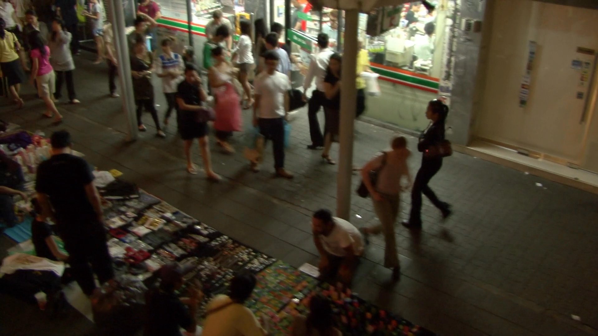 Pedestrians navigate a vibrant night market located along a riverfront in Bangkok. The footage captures the transition from dusk to evening as crowds shop among various stalls and surrounding city traffic flows.