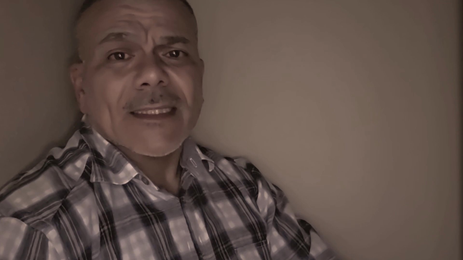 A man in a plaid shirt speaks directly to the camera in a serious and engaged manner.