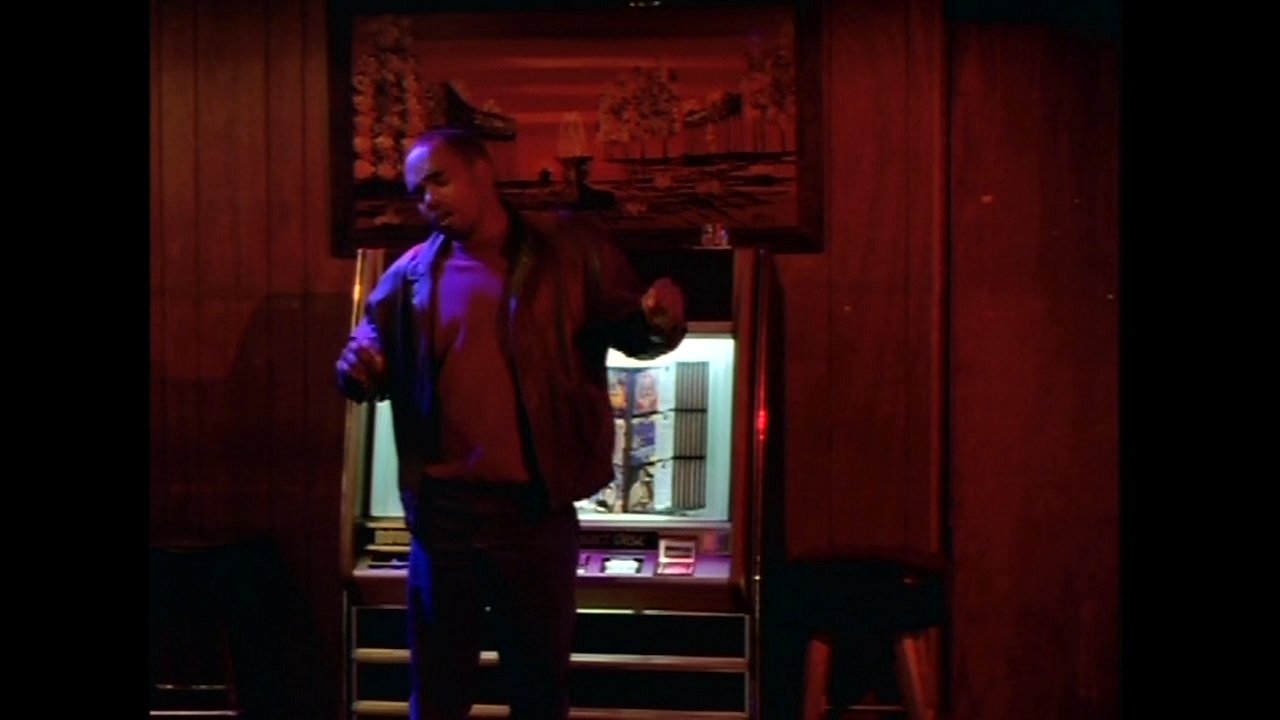 A man dances rhythmically in front of a glowing jukebox within a dimly lit, atmospheric lounge setting.