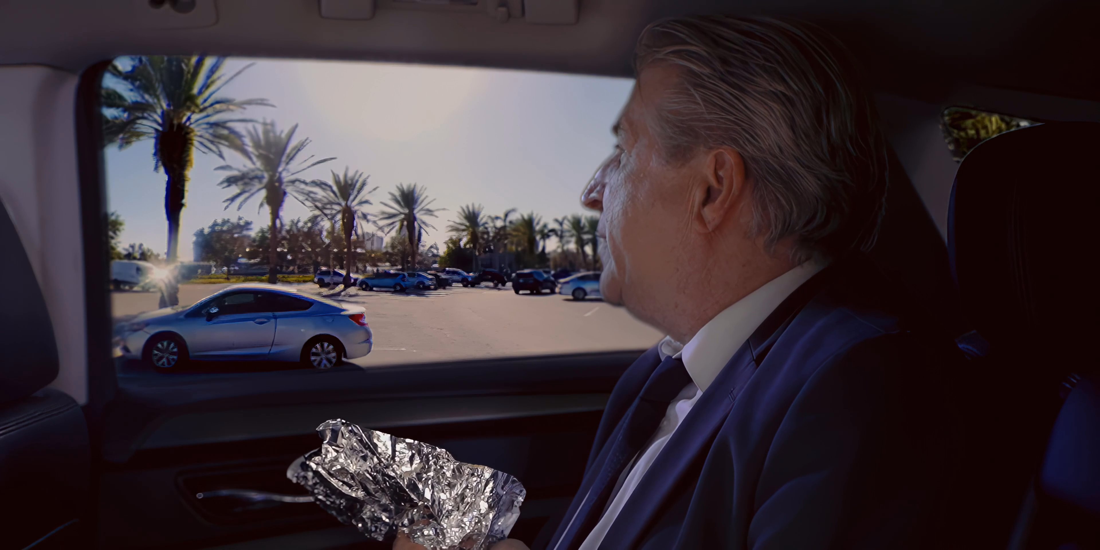 Two professional men sit in a parked vehicle, engaging in a focused conversation while one prepares to eat a wrapped meal.