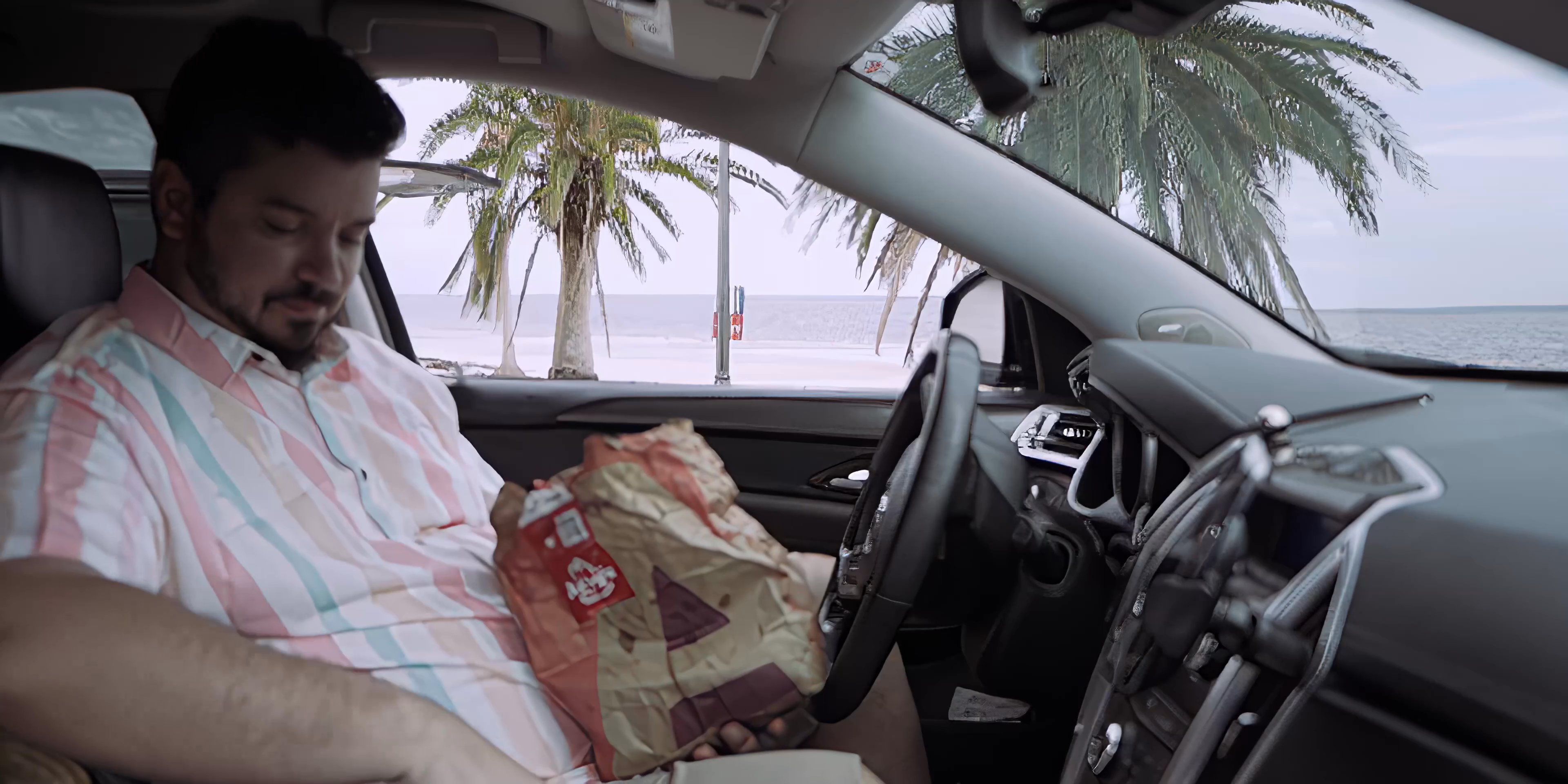 A man sits inside a parked car overlooking the coast and opens a brown paper bag to consume a meal.