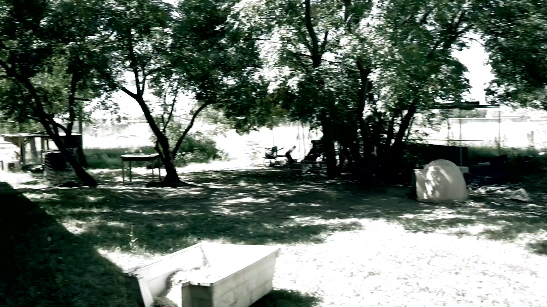 A residential property features a collection of outdoor fitness equipment placed in a shaded backyard area. The camera moves through the space, providing a panoramic view of the training equipment set amongst mature trees.