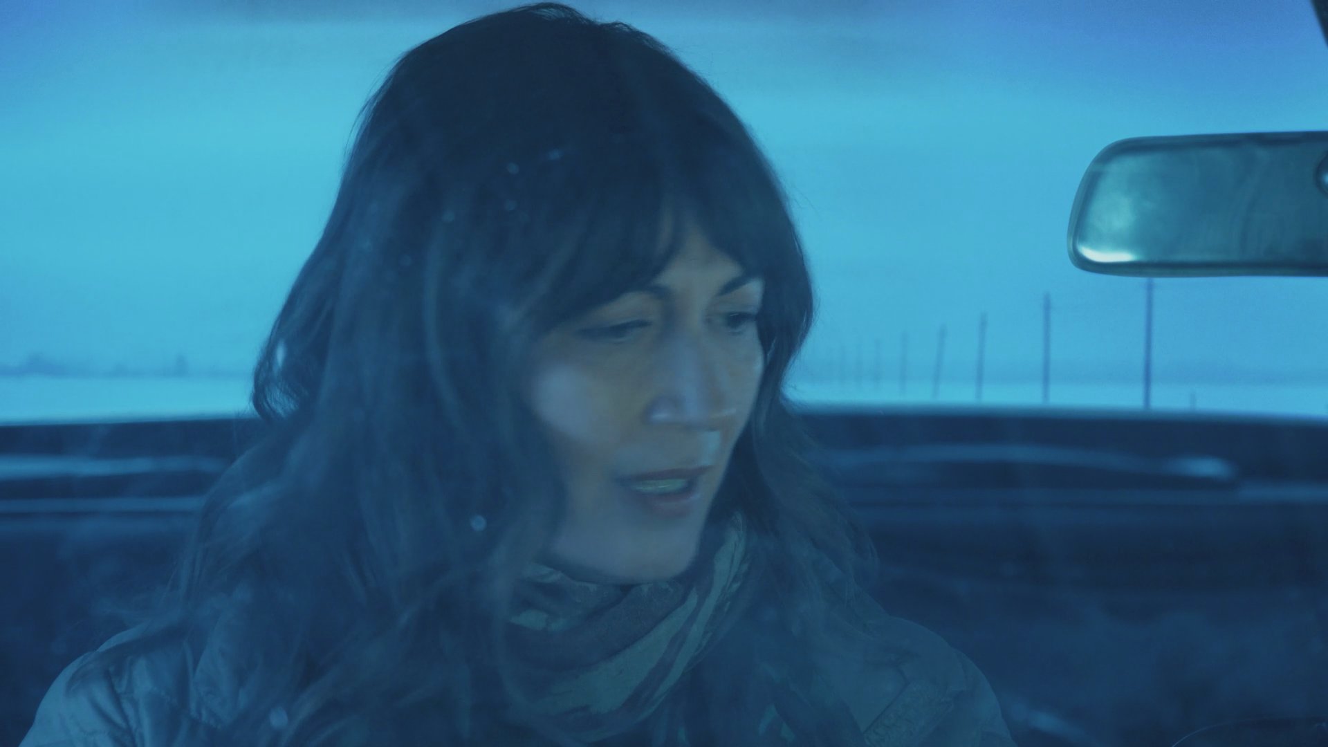 A woman hands cash to a man while seated inside a moving vehicle. The pair engage in an intense discussion during their journey through a cold, wintry landscape.