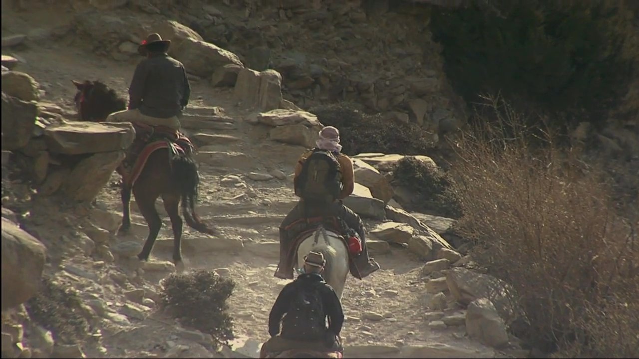 Travelers navigate a rugged, high-altitude mountain landscape on horseback toward a traditional stone monastery and colorful chorten. This expedition highlights remote transit through the stark, mountainous terrain of the Mustang region.