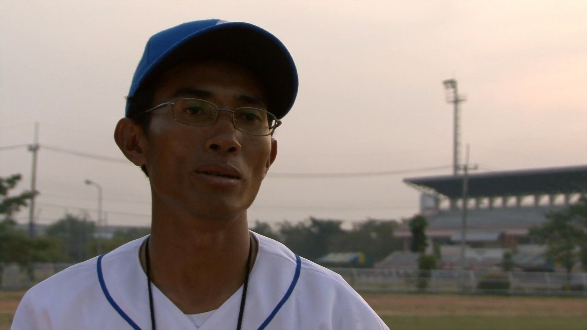 A baseball coach wearing a team uniform and cap stands on a field and speaks to an interviewer. The setting is an outdoor stadium during the evening.
