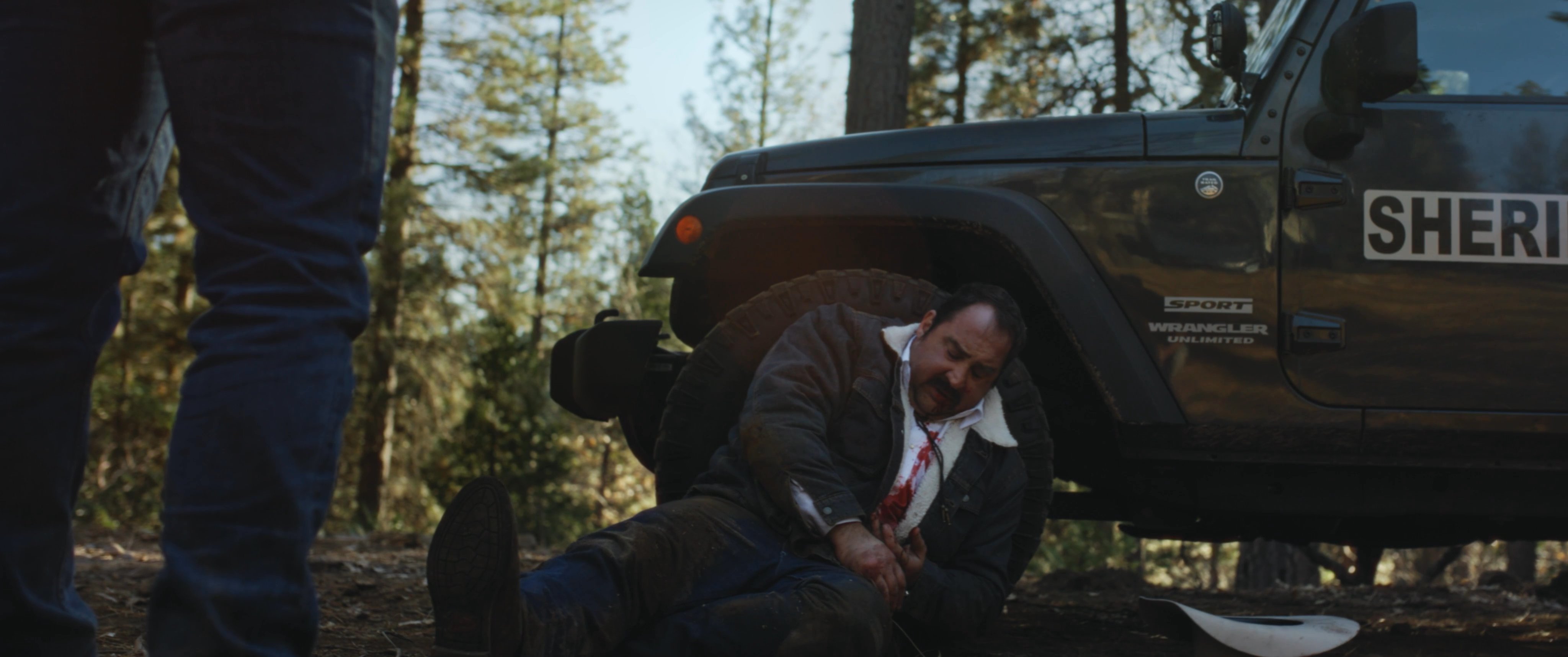 An injured sheriff in uniform sits on the forest floor beside a department vehicle while another individual approaches him. The scene captures a moment of high tension and physical distress.