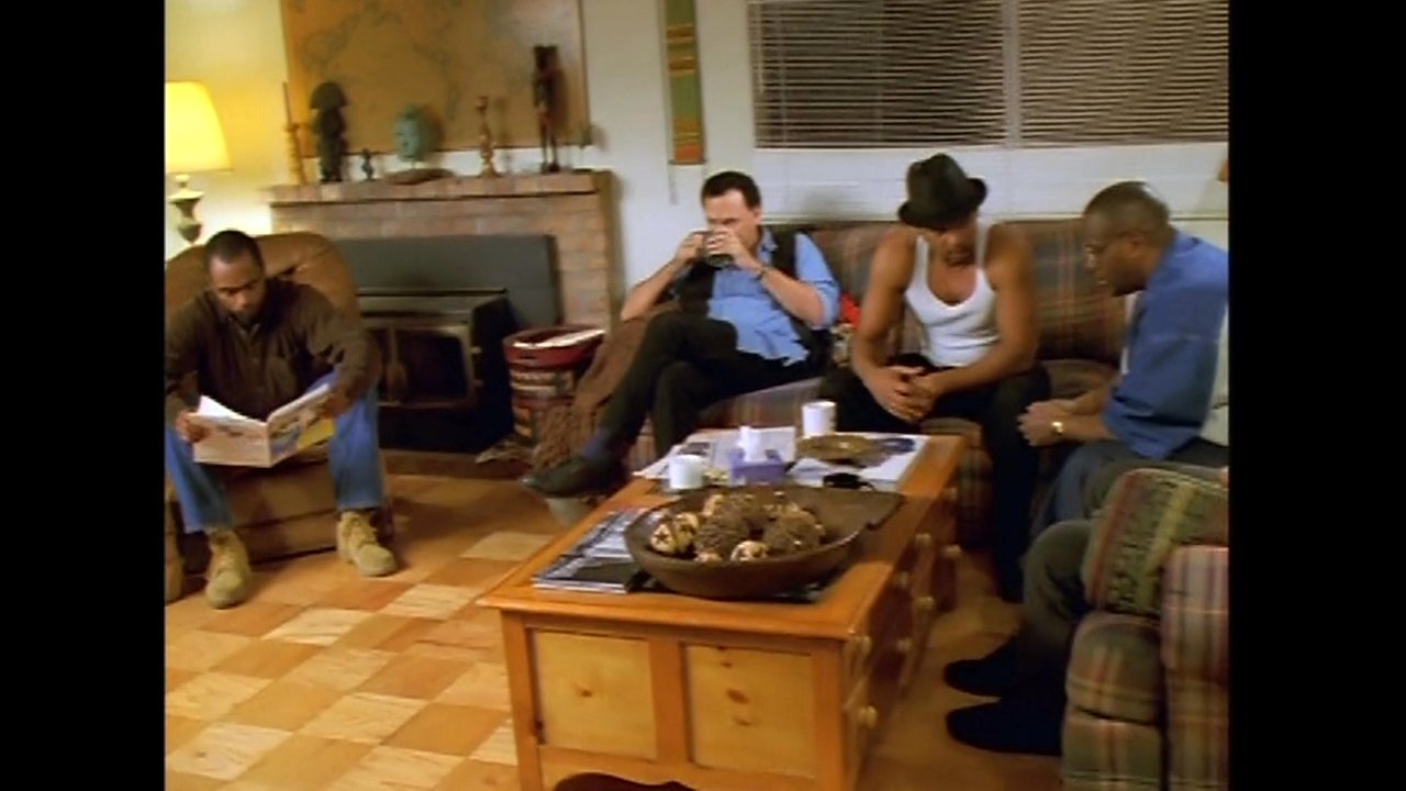 A couple embraces by a marsh at sunset, transitioning to a group of men conversing in a residential living room setting.