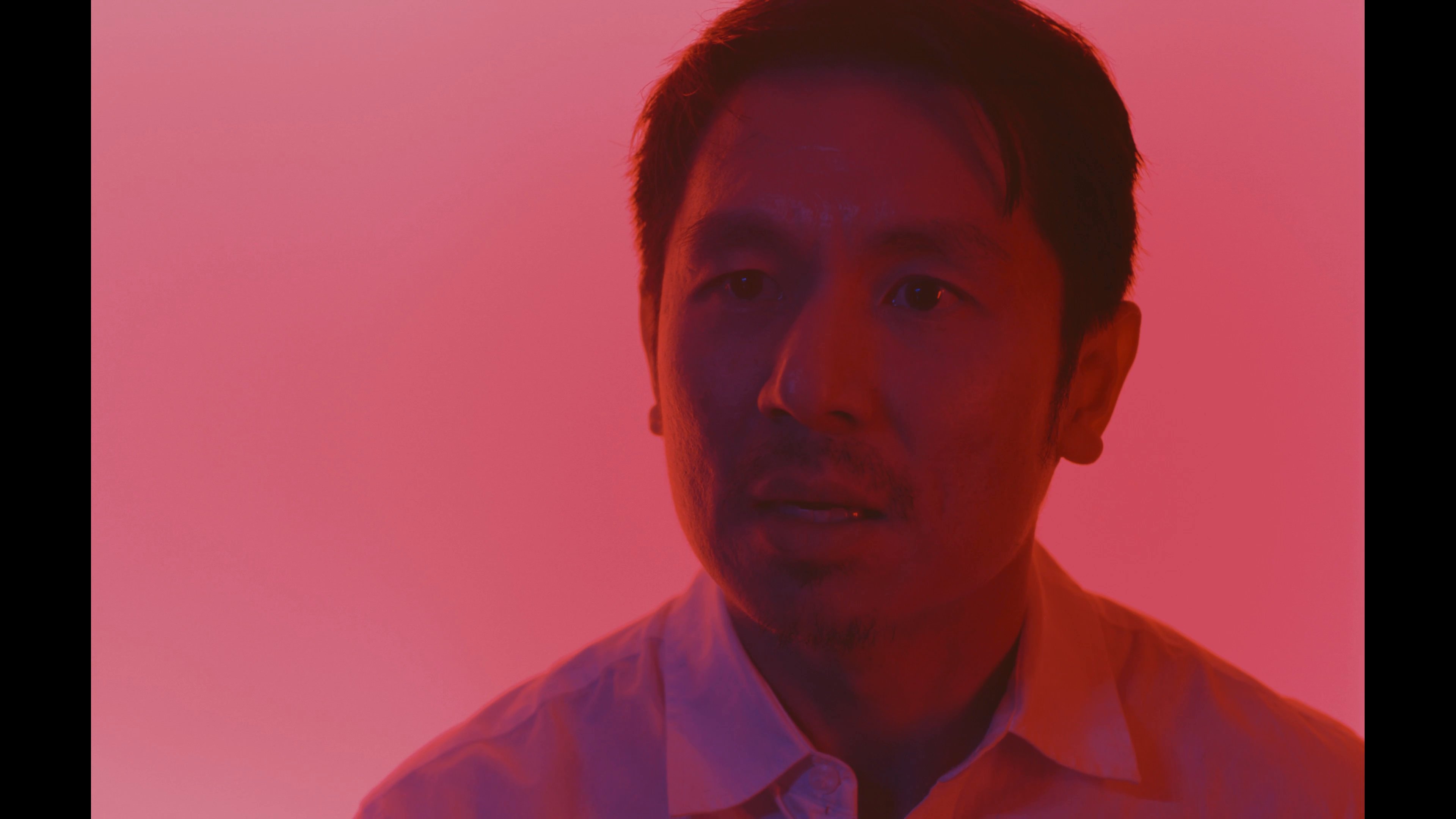 Two individuals engage in a serious, emotionally charged conversation while surrounded by saturated red lighting. The scene focuses on their subtle facial expressions and reactive body language during the exchange.