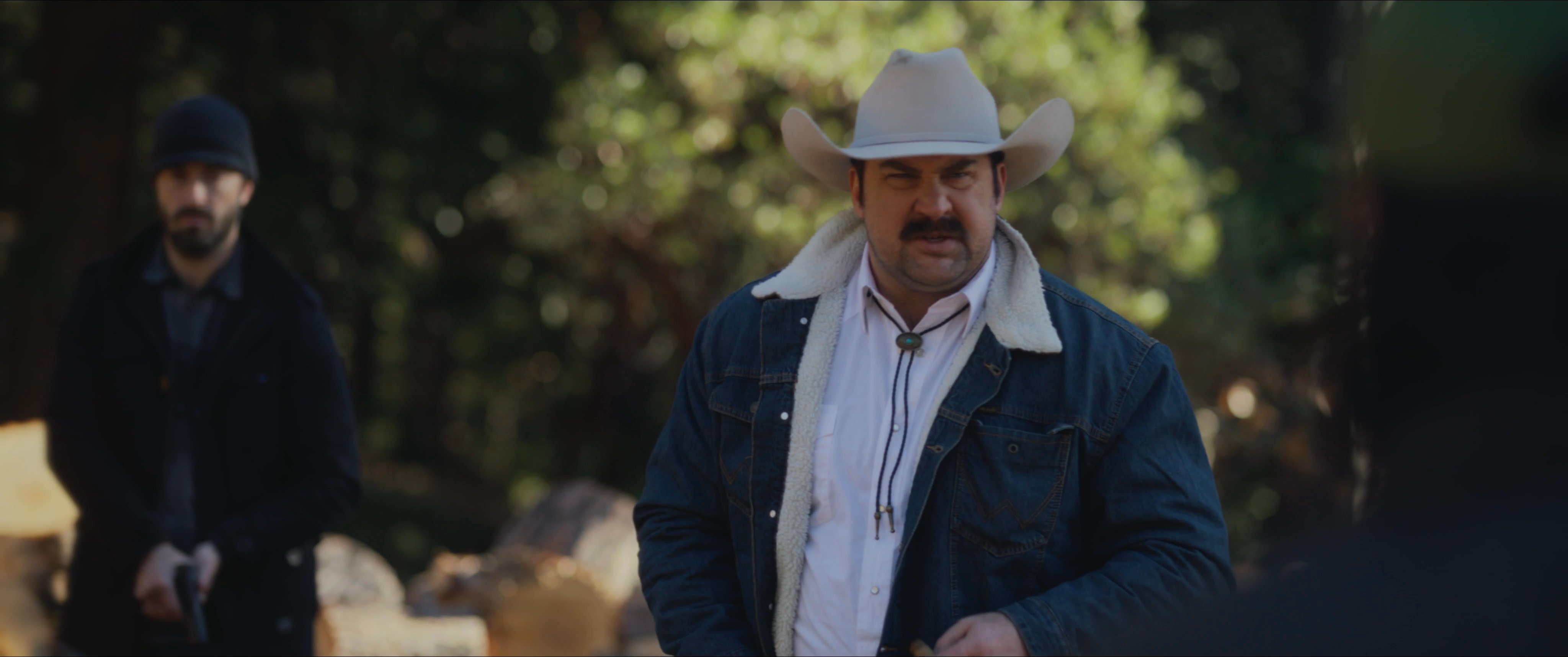 A man in a cowboy hat engages in a tense standoff with a sniper positioned in a forest setting. The confrontation escalates as participants display firearms in a high-stakes encounter.
