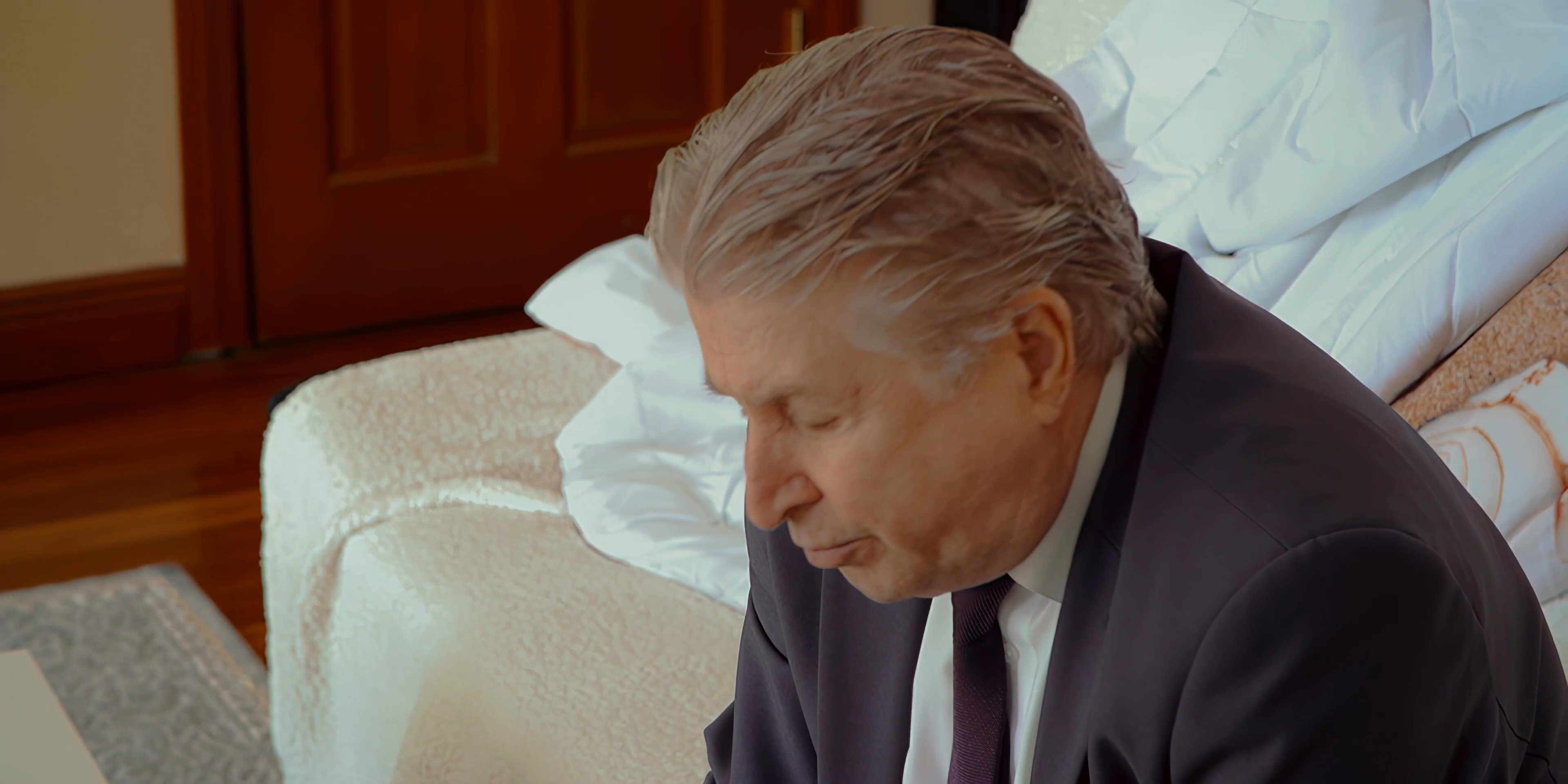 A man in a red shirt expresses frustration while engaged in a tense conversation with an older man sitting on a bed. The interaction conveys significant interpersonal strain and professional discord.