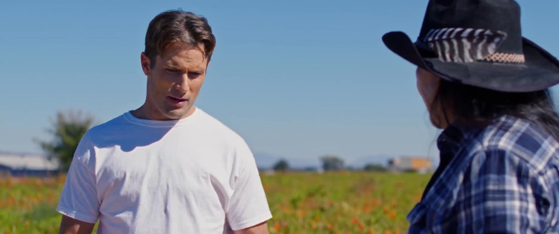 An indigenous farmer explains agricultural processes to a visitor while standing in a sprawling chili field. They engage in a focused discussion regarding cultivation and harvesting techniques in a desert landscape.