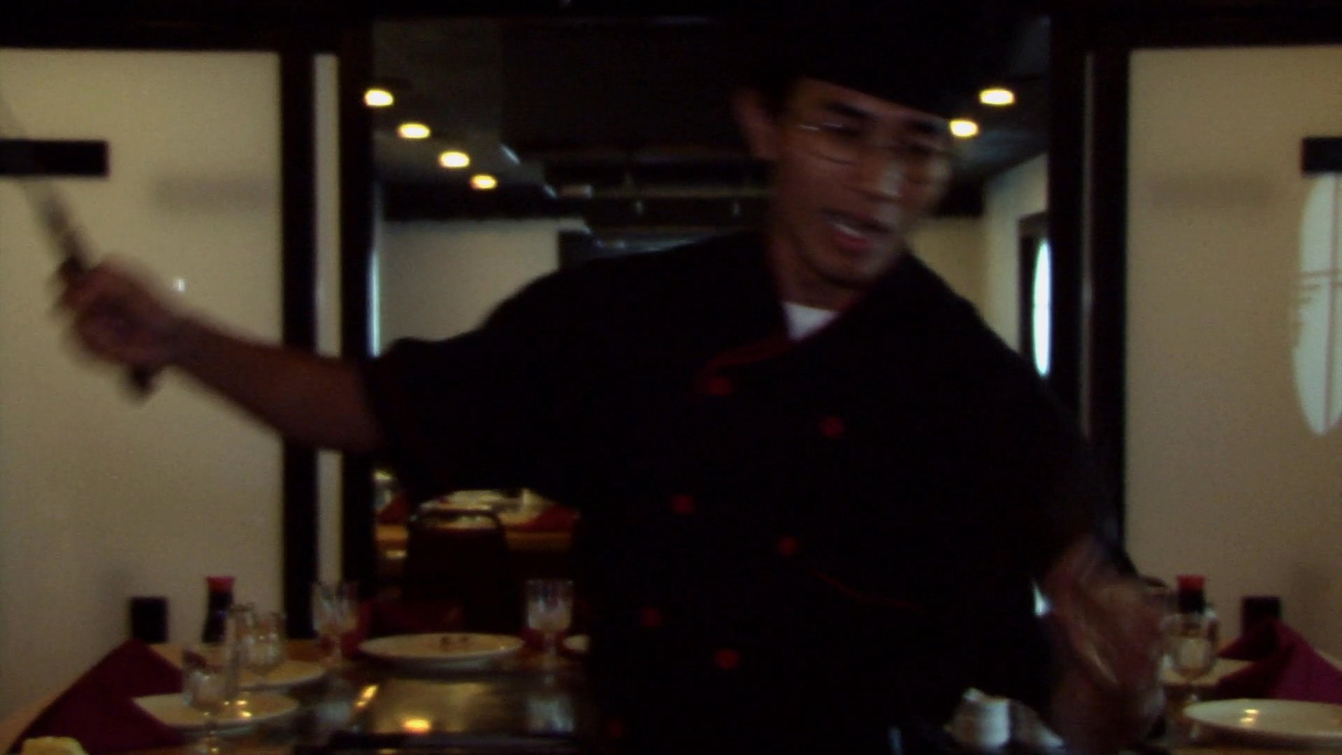 A skilled chef prepares a meal on a teppanyaki grill, executing technical cooking maneuvers and a dramatic flaming technique to finish the dish.