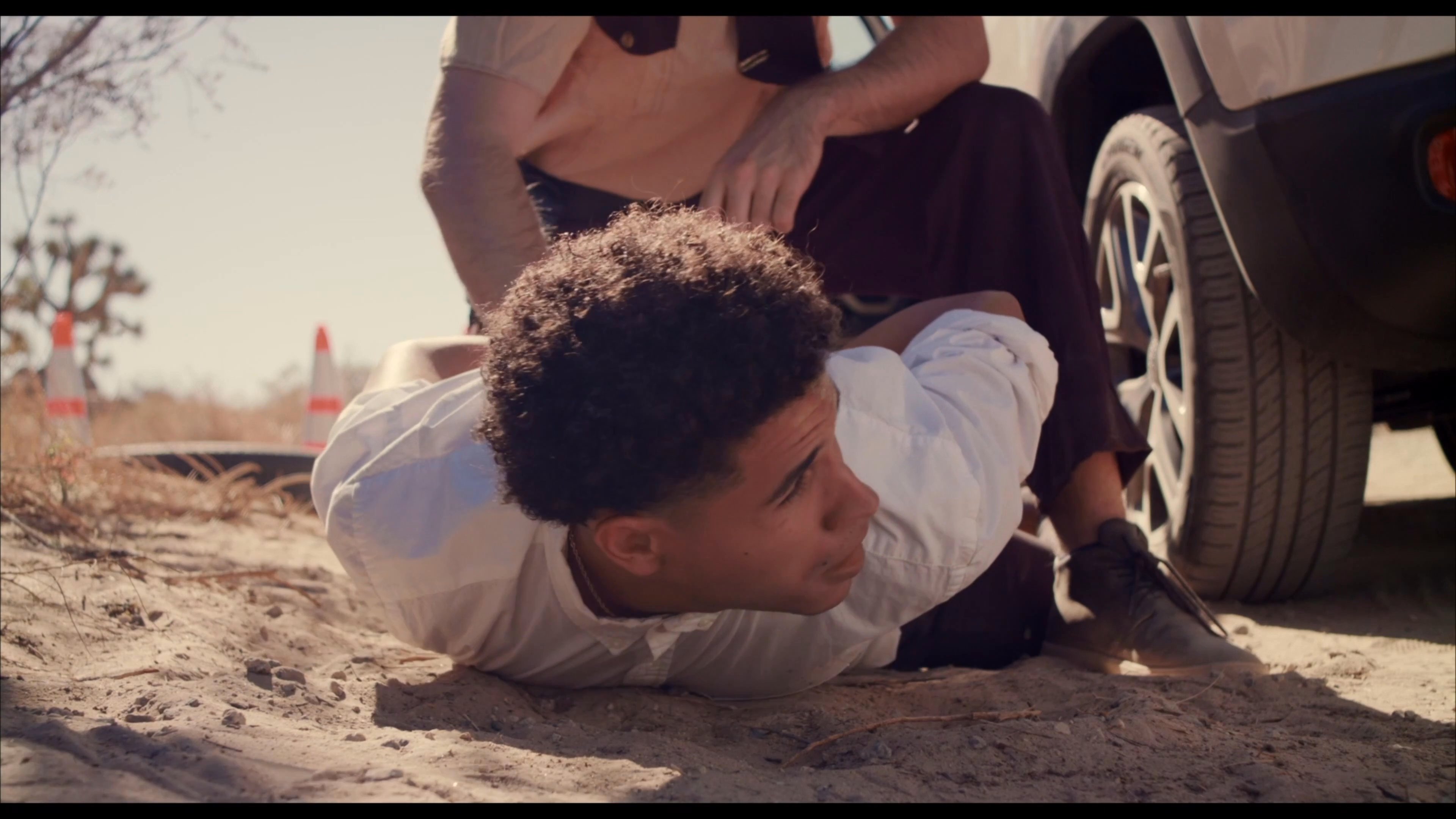 A DEA agent executes an arrest in a desert landscape as a suspect is restrained on the ground. The agent maintains control of the scene while surveying the surrounding environment.