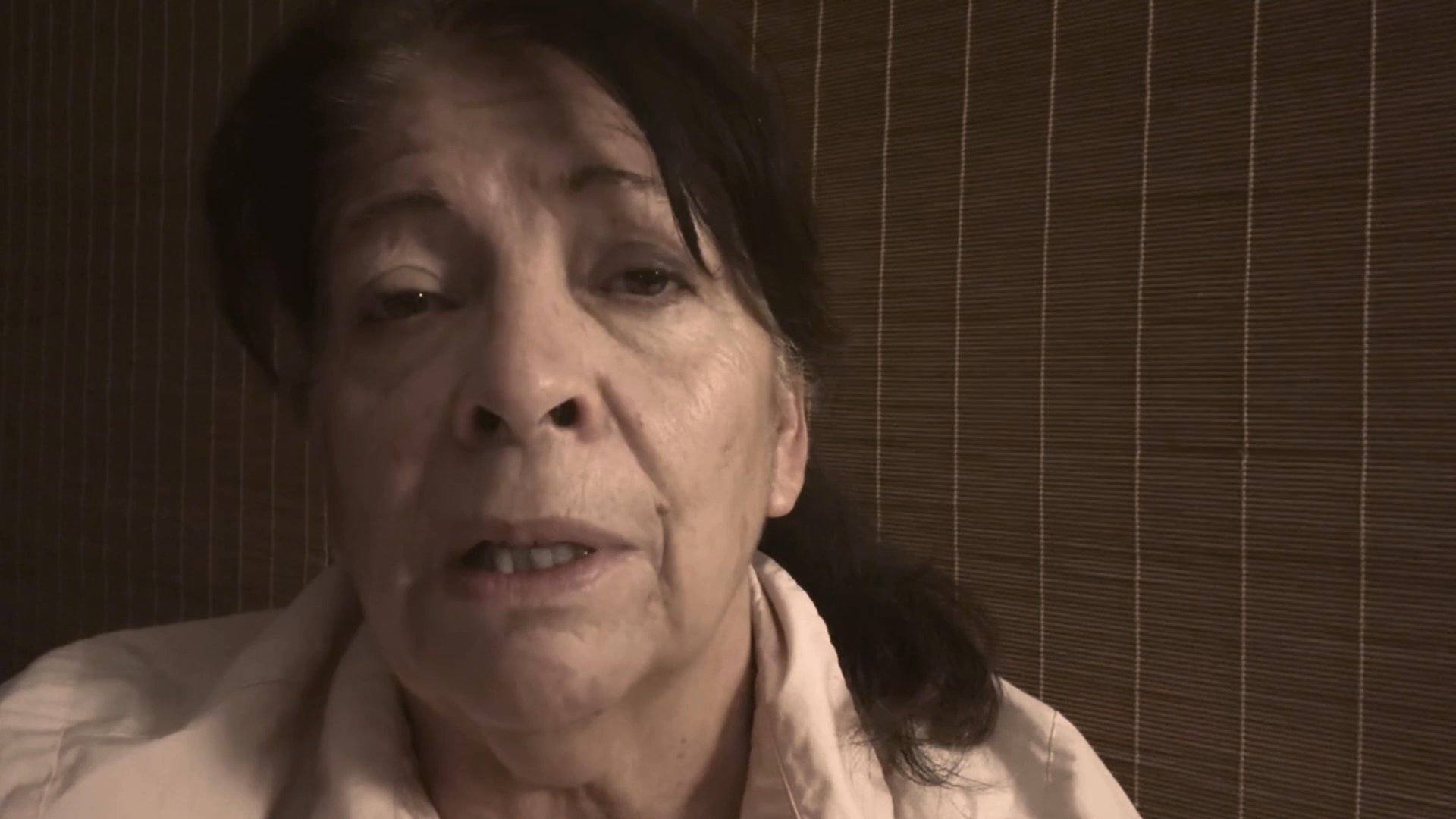 An older woman speaks directly to the camera with a serious and reflective expression. She maintains a consistent, steady focus while engaged in a thoughtful monologue.