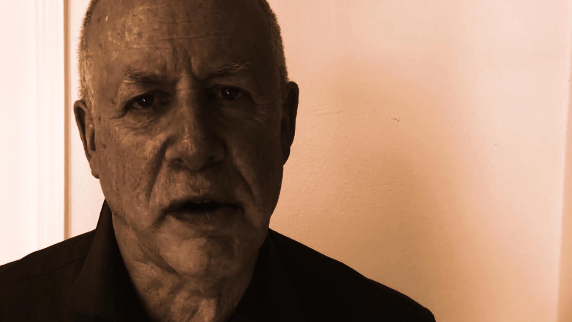 An older man delivers a serious message while looking directly into the camera in a professional setting.