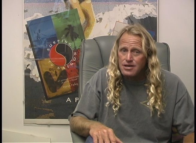 A man with long blonde hair discusses surfing while sitting in an office environment. He utilizes hand gestures to emphasize his points during the interview.
