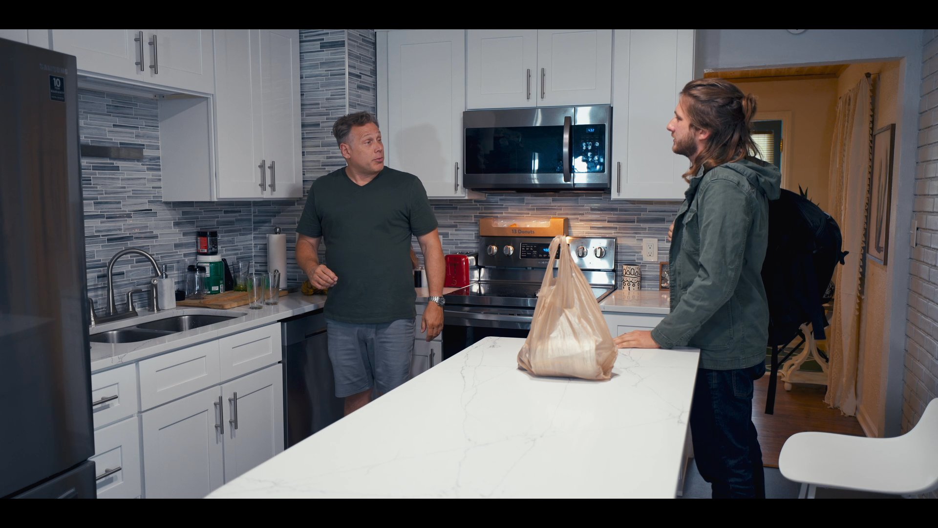 Two men enter a modern residential kitchen, where one person proceeds to unpack a grocery bag onto the central island while the other observes.