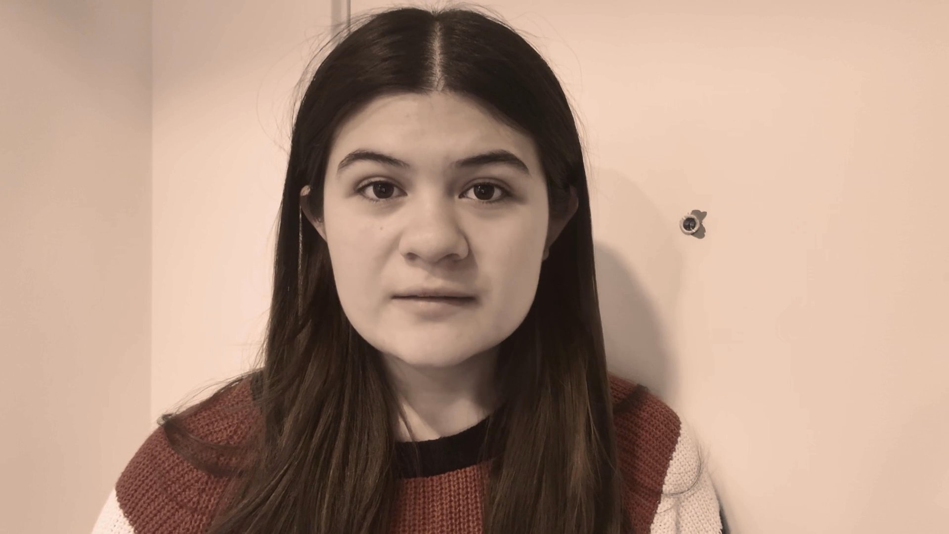 A young woman speaks directly to the camera while positioned in a bedroom with sheer curtains billowing in the breeze from an open window.