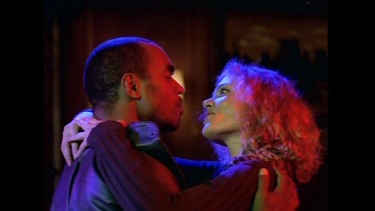 A man and a woman share a close, affectionate moment in a dimly lit, moody environment. Their intimate interaction highlights a deep personal connection as they remain locked in a slow, tender embrace.