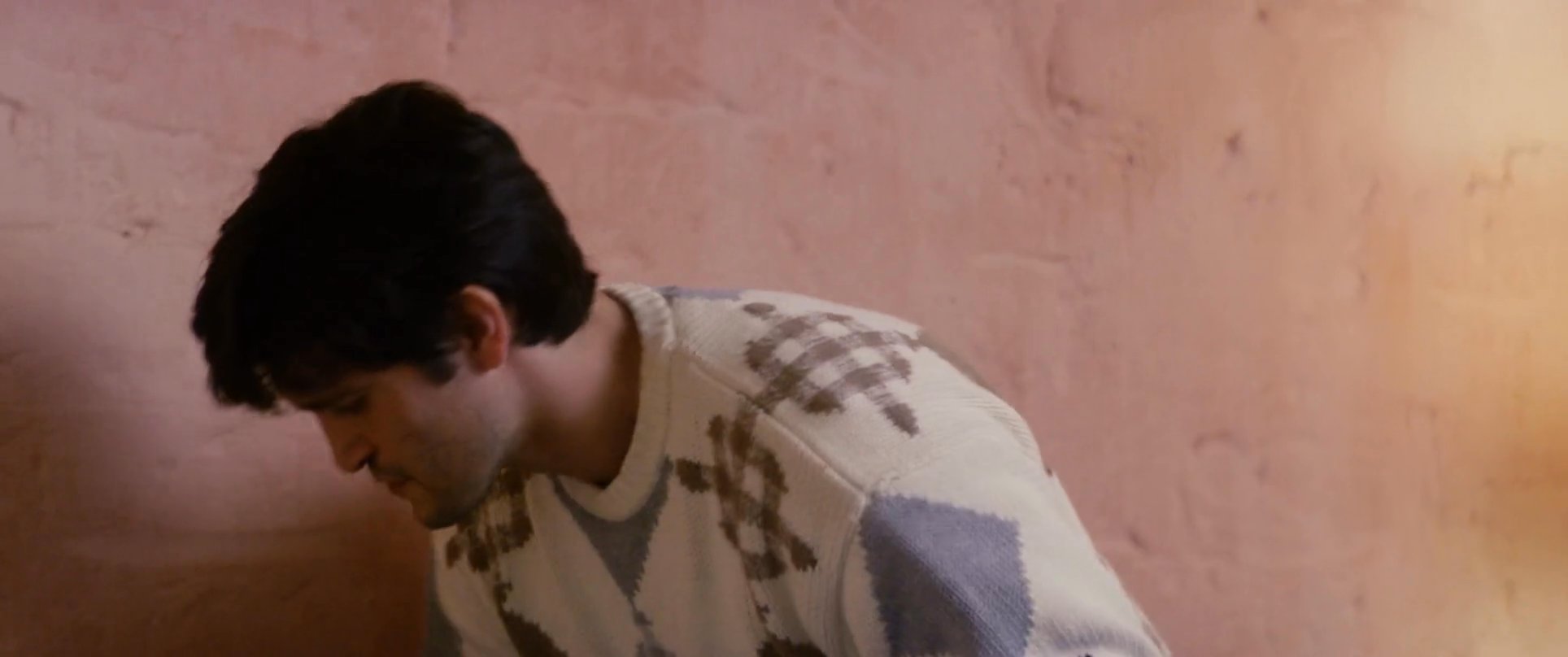 A man provides comfort and tender care to another man lying on a bed. The intimate interaction takes place in a dimly lit, private setting.