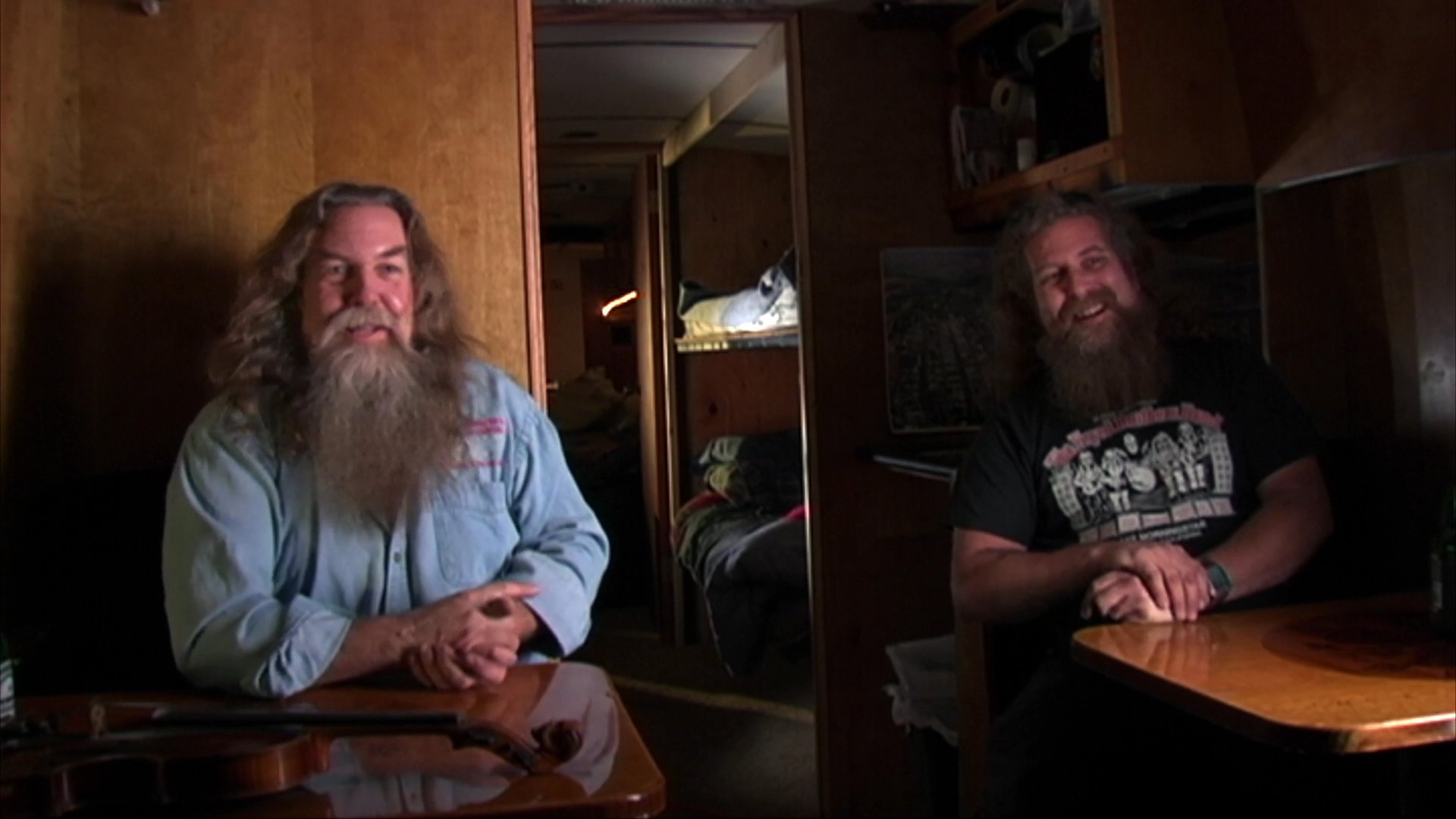 A long-haired man with a beard sits at a table in a dimly lit room, sharing personal anecdotes and reflections during an interview. He uses expressive hand gestures to emphasize points throughout his conversation.