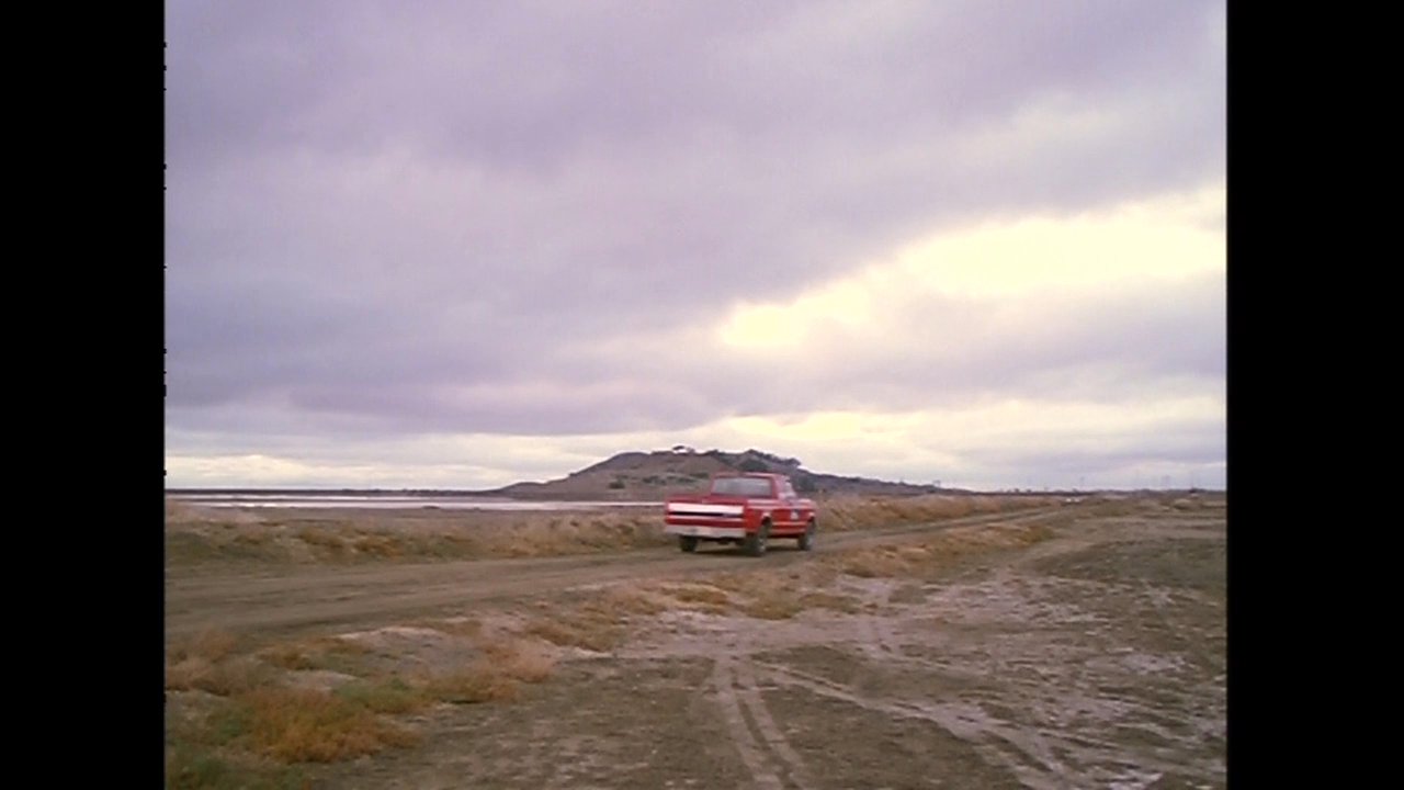 A man and woman travel in a vehicle through an expansive, industrial landscape characterized by large salt mounds and water basins.