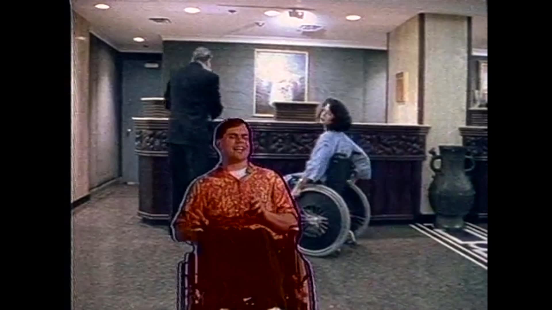 A man in a suit engages in a professional dialogue with a woman seated in a wheelchair at a lobby reception desk. The scene captures a focused exchange between the two individuals in a formal office setting.
