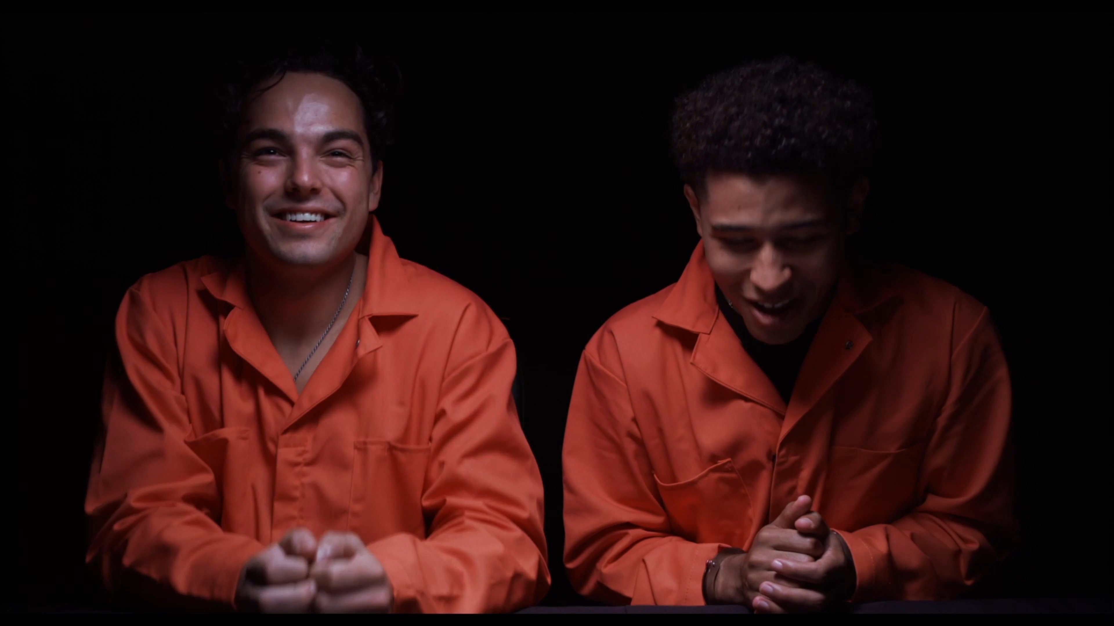 Two individuals in orange prison jumpsuits and handcuffs participate in a humorous interrogation session with law enforcement officials.