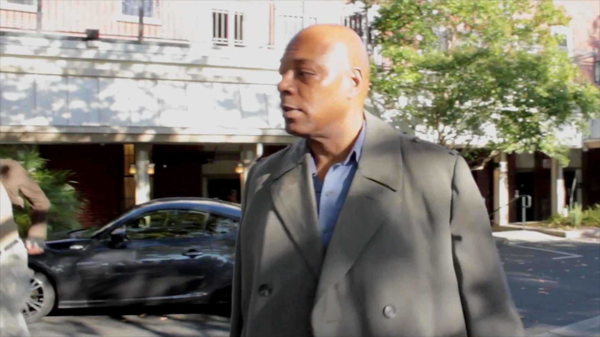 A blonde woman watches from the sidelines as two men meet on an urban street to engage in a focused, in-person conversation.