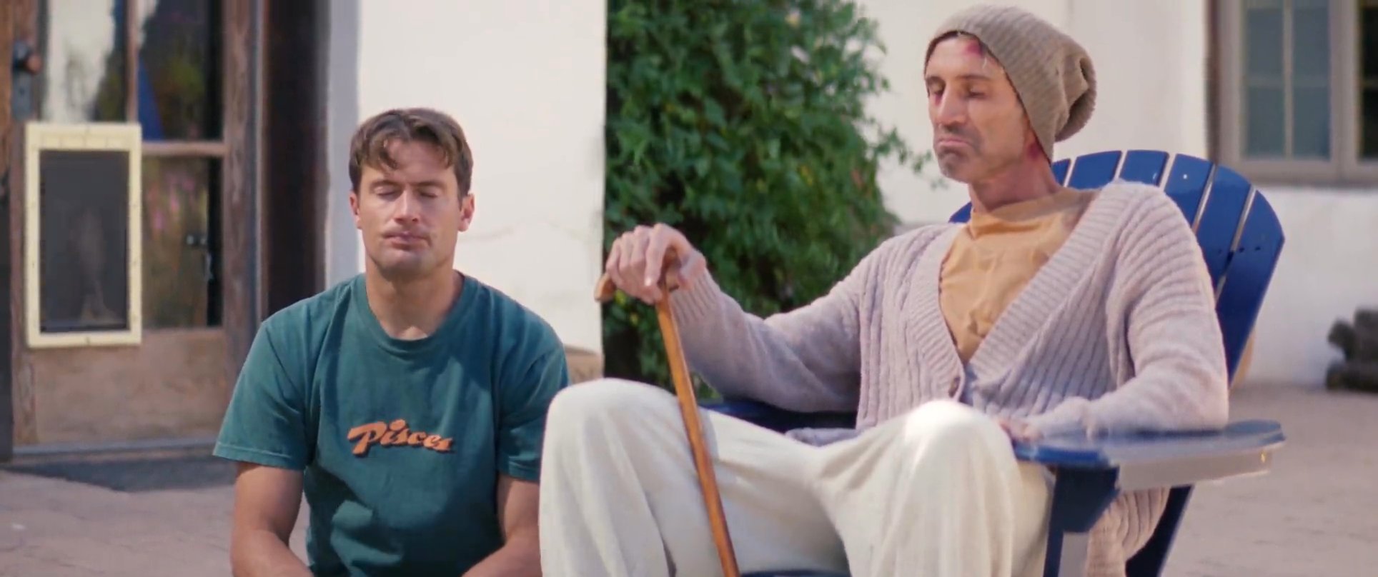 An older man in a knitted cardigan and beanie engages in a thoughtful conversation with a younger man on an outdoor patio set against a rural landscape.