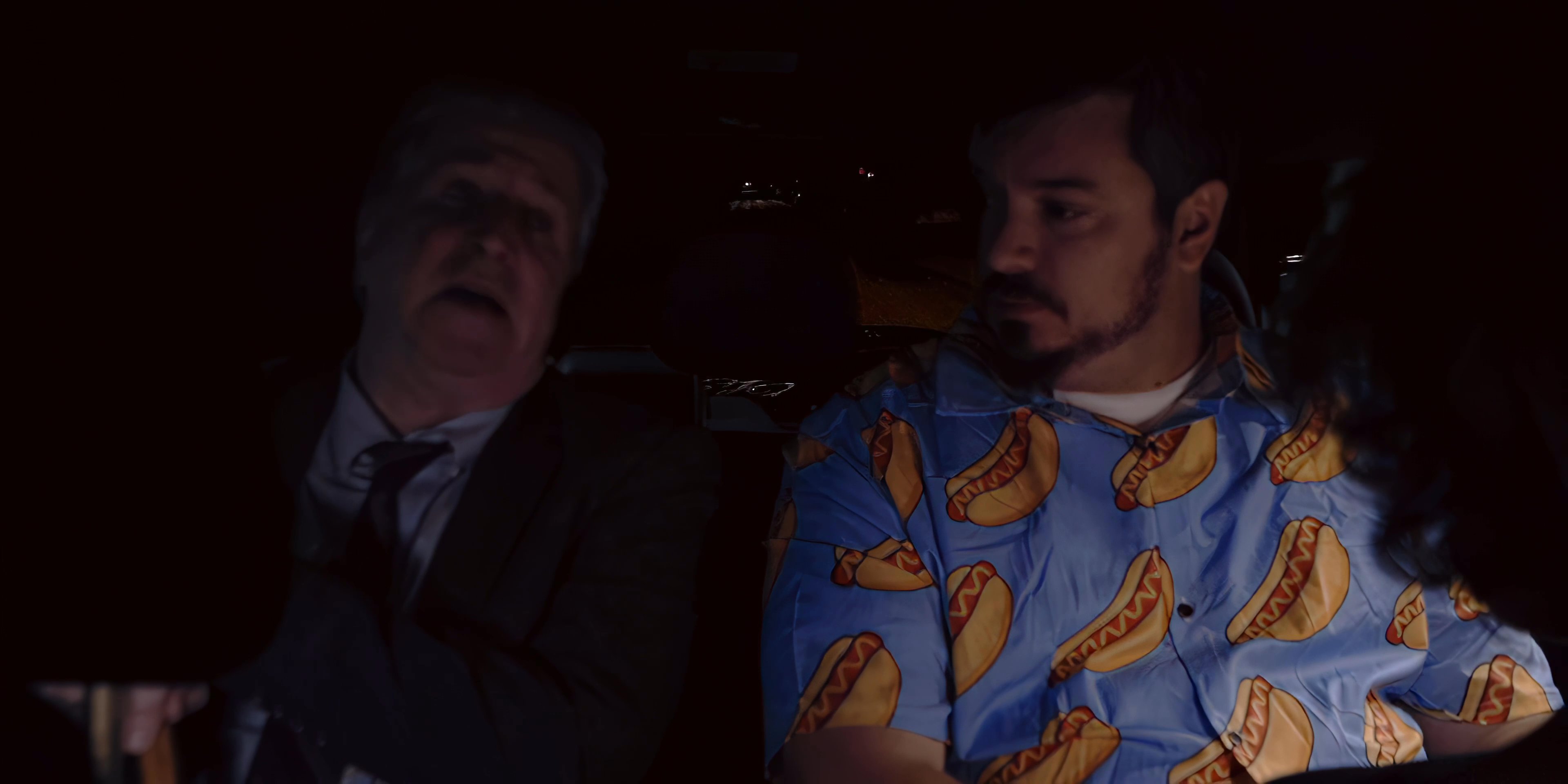 Two men sit inside a vehicle at night, engaging in a quiet conversation while traveling through an illuminated city environment.