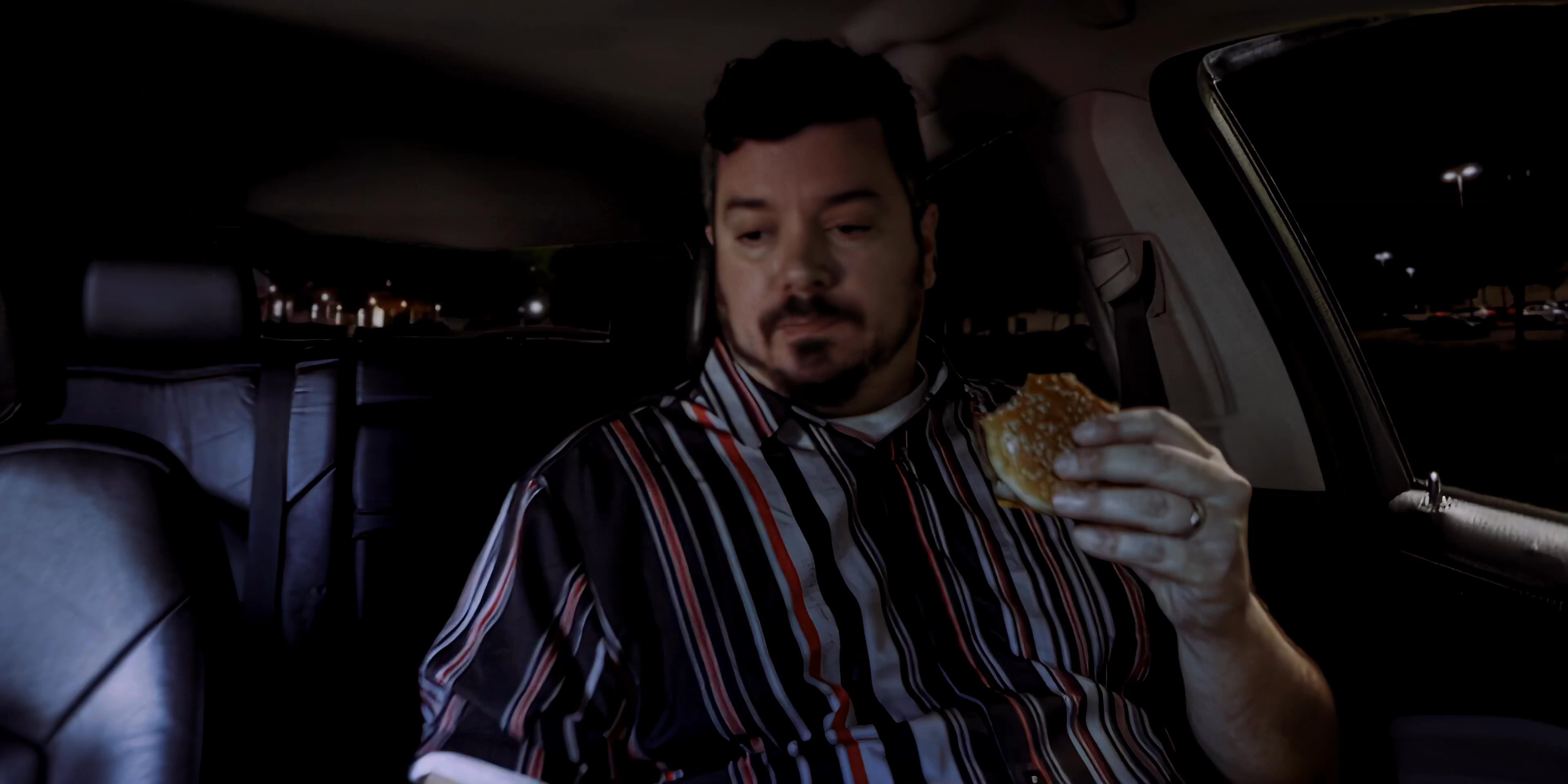 A man sits alone in his vehicle in a dark parking lot at night, eating a hamburger while receiving an incoming call on his smartphone.