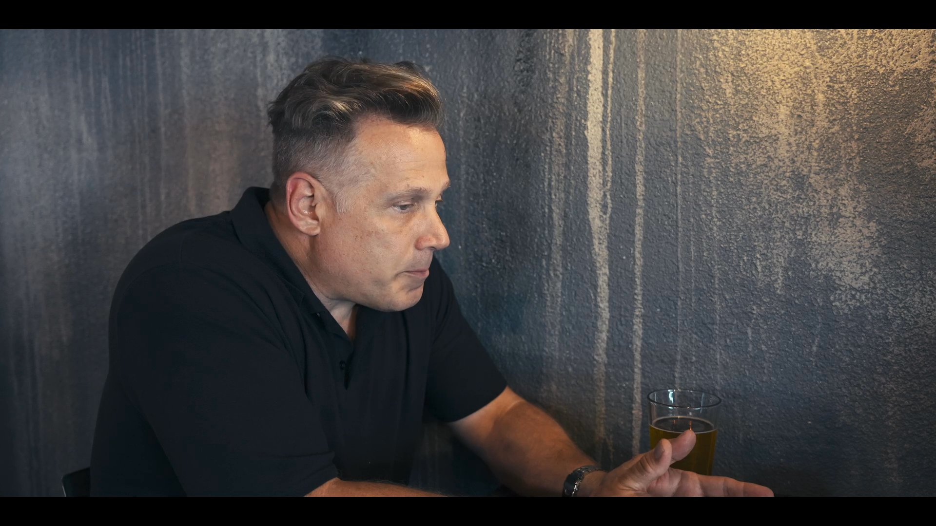 A man in a dark polo shirt engages in an active, expressive conversation while seated before a textured background. He uses hand gestures to emphasize his points during the interview session.