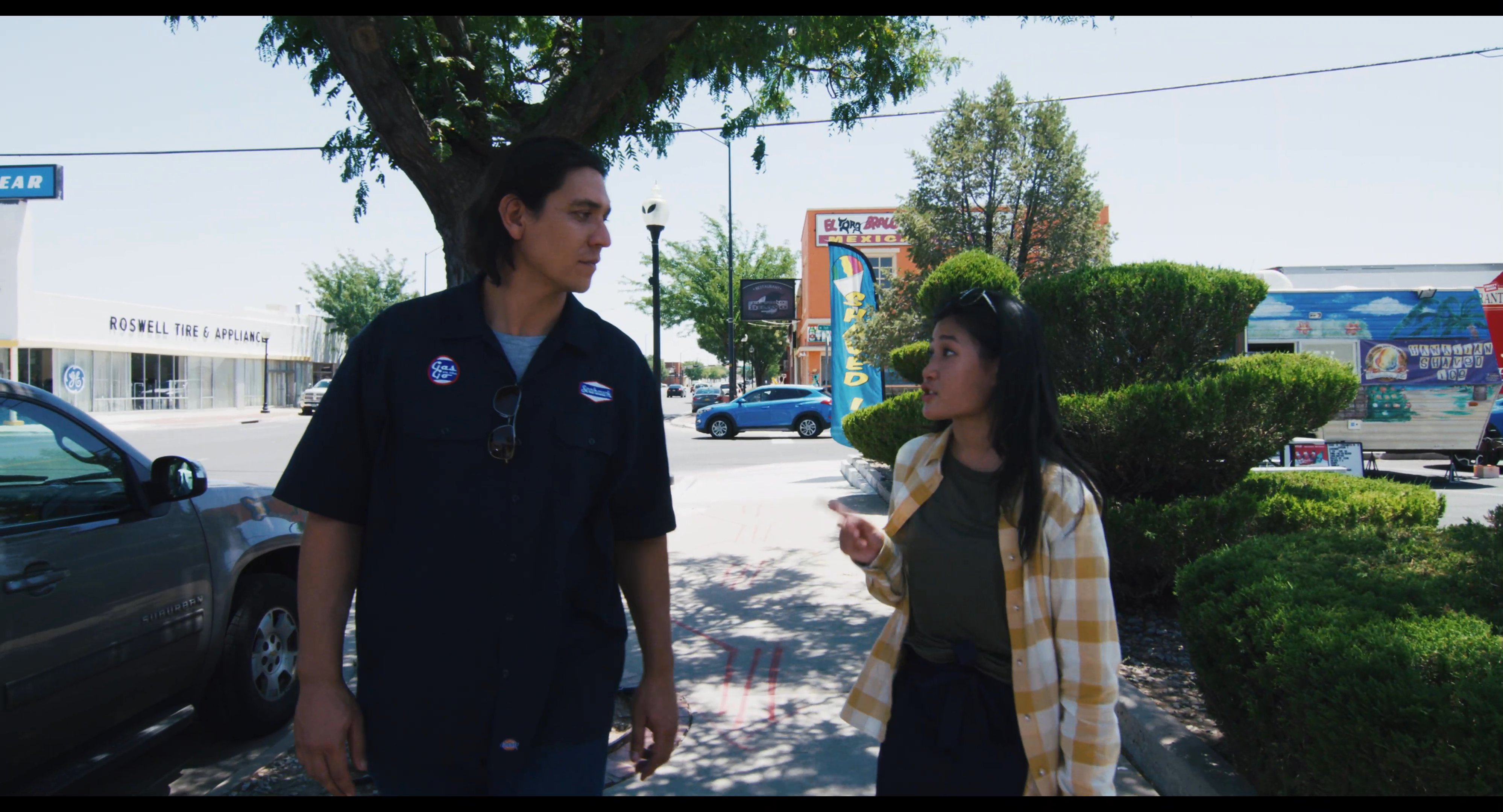 A man and woman walk together while engaged in conversation outside of a retail shopping complex. They traverse a pedestrian area adjacent to a parking lot during a bright day.