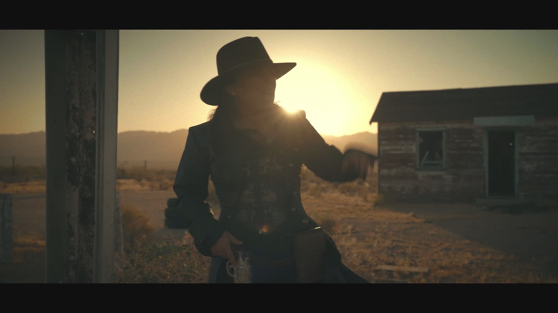 At sunset on a weathered frontier homestead, a hat-wearing woman approaches a long-haired figure suspended by a rope beneath a timber structure. The tense encounter ends in a close-up of hands gripping near the figure’s neck.