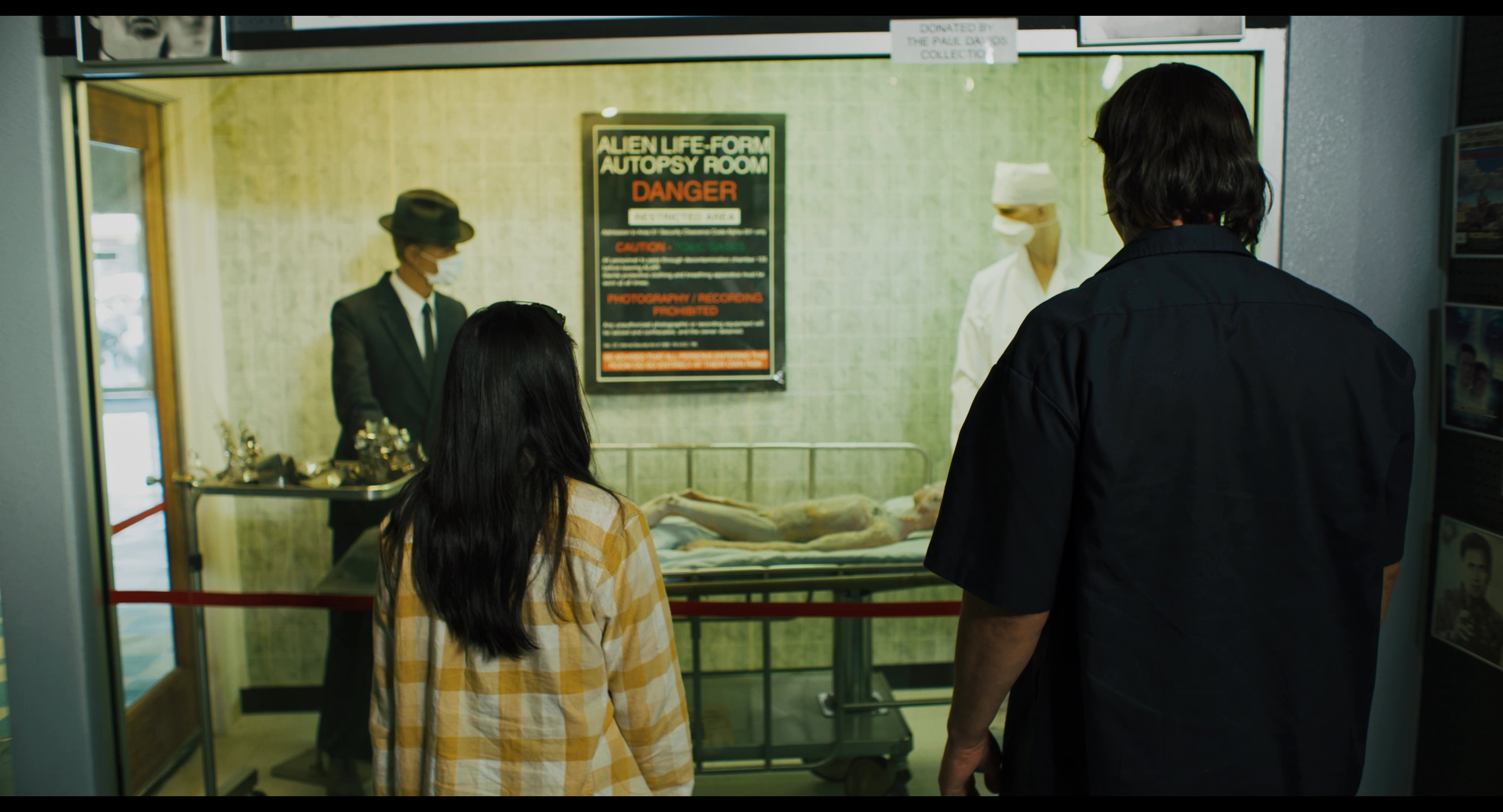 Two visitors explore a themed museum featuring alien-related displays, including life-sized extraterrestrial figures and a staged autopsy exhibit. They observe historical and fictional artifacts that characterize the pop culture portrayal of UFO encounters.
