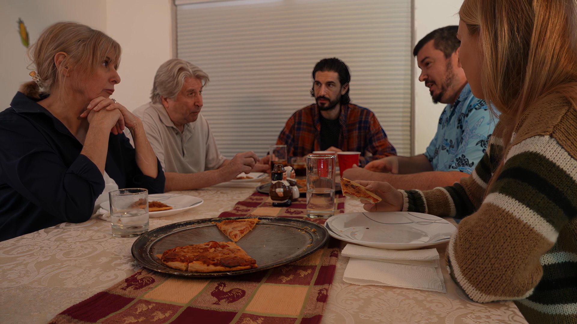 A diverse group of adults enjoys a shared meal and casual conversation around a dining table. The scene highlights a communal social environment centered on food and interaction.