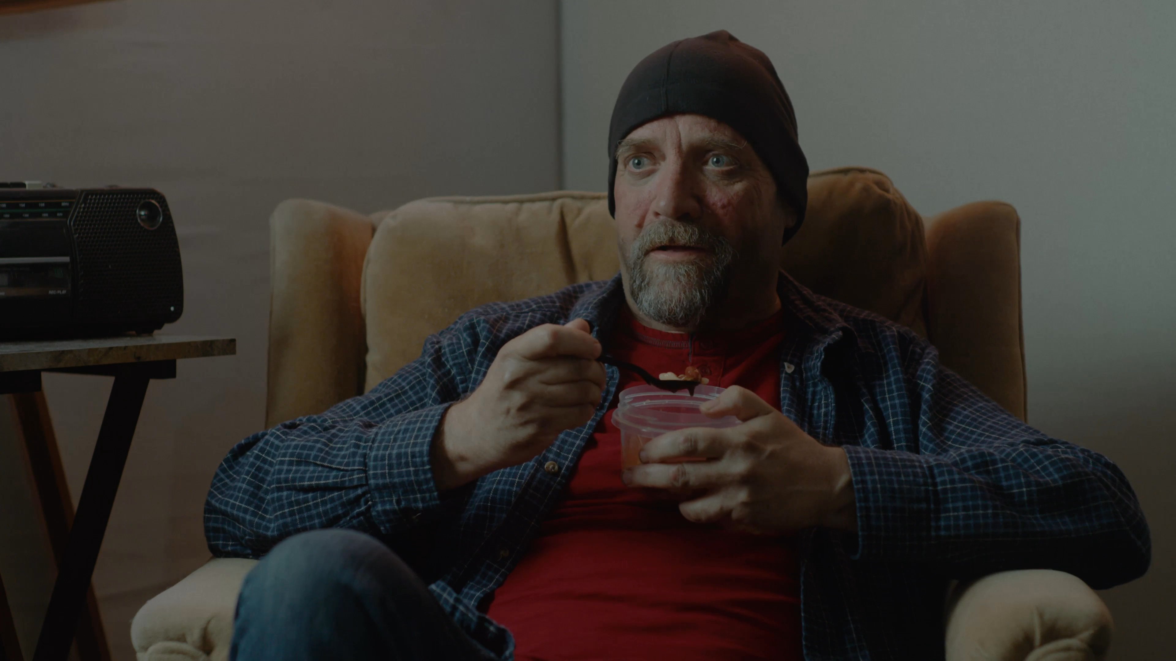 Two men engage in a conversation while sitting in a living room, with one man eating a snack from a container while the other speaks.